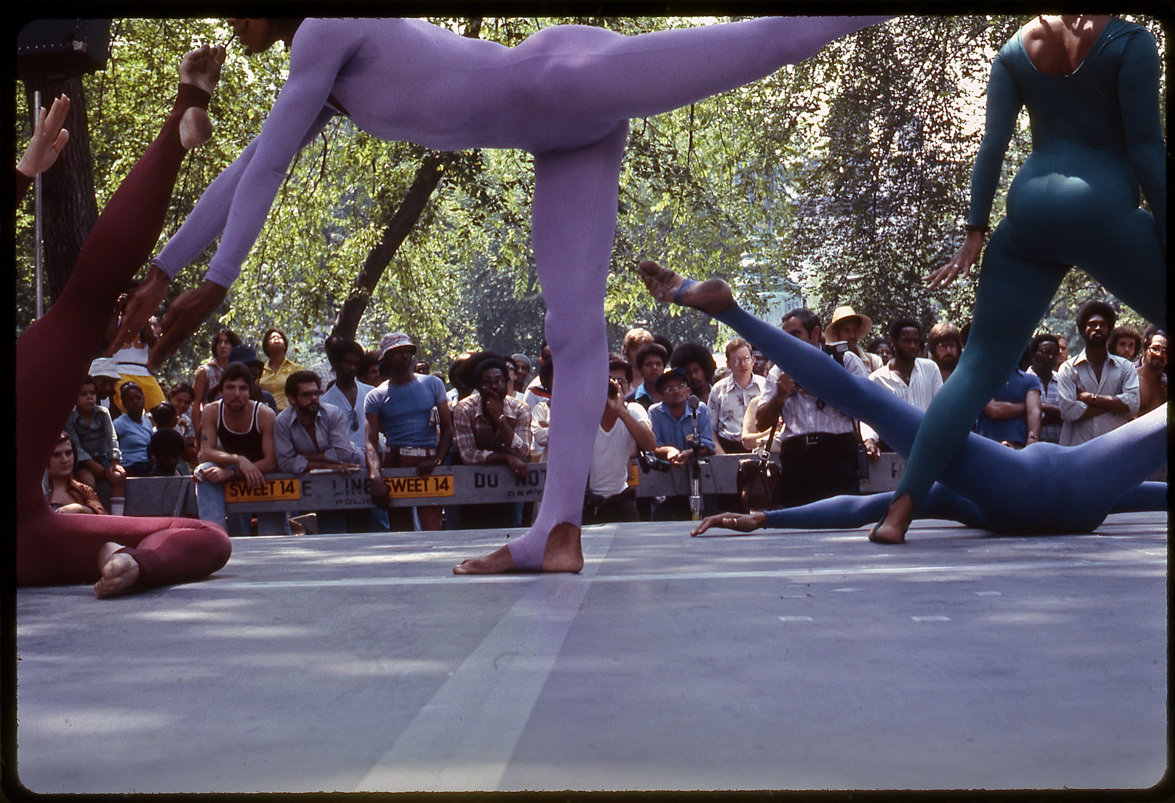 Street level view of dancers performing in a park, with a crowd of people viewing the performance in the background underneath a canopy of trees. At center is a dancer wearing a lavender bodysuit, standing on one leg with their other leg raised behind them and their arms pointed down to the ground. Other dancers, in burgundy, blue, and teal bodysuits, are in various dynamic positions surrounding the central figure. 