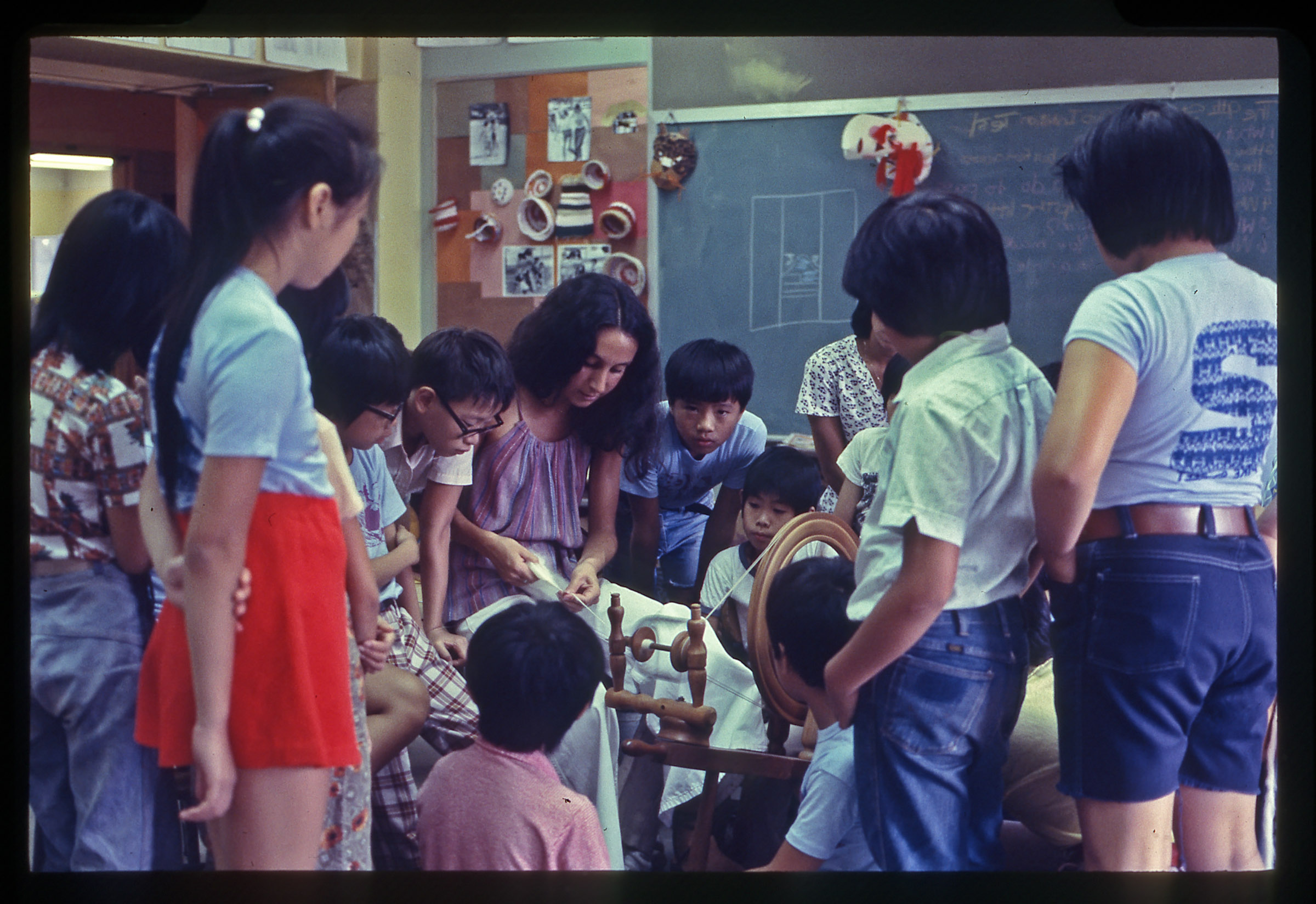 A woman with long dark hair parted down the middle is surrounded by over a dozen grade school children in a classroom. There is a chalkboard and various crafts on the wall in the background. Everyone's focus is toward the center of the image, where the woman is demonstrating weaving using white yarn and a manual wooden device.