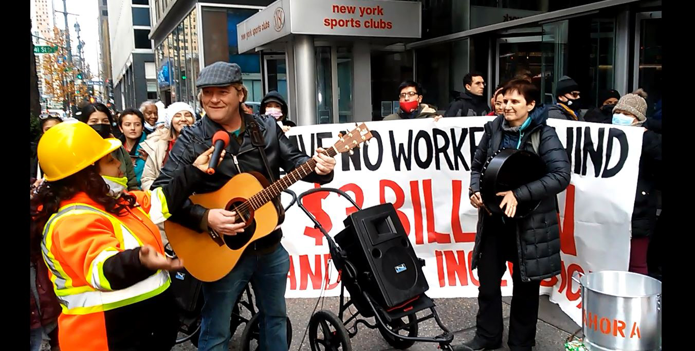 Photograph of a smiling person playing an acoustic guitar next to a portable speaker. There is someone holding a circular case on one side of the guitarist and...