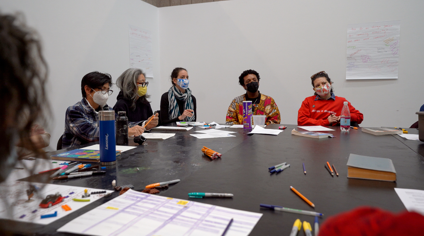 A group of people sit around a rectangular table in a windowless interior. Pencils, markers, papers, and books are scattered across the table. Five people sit directly...