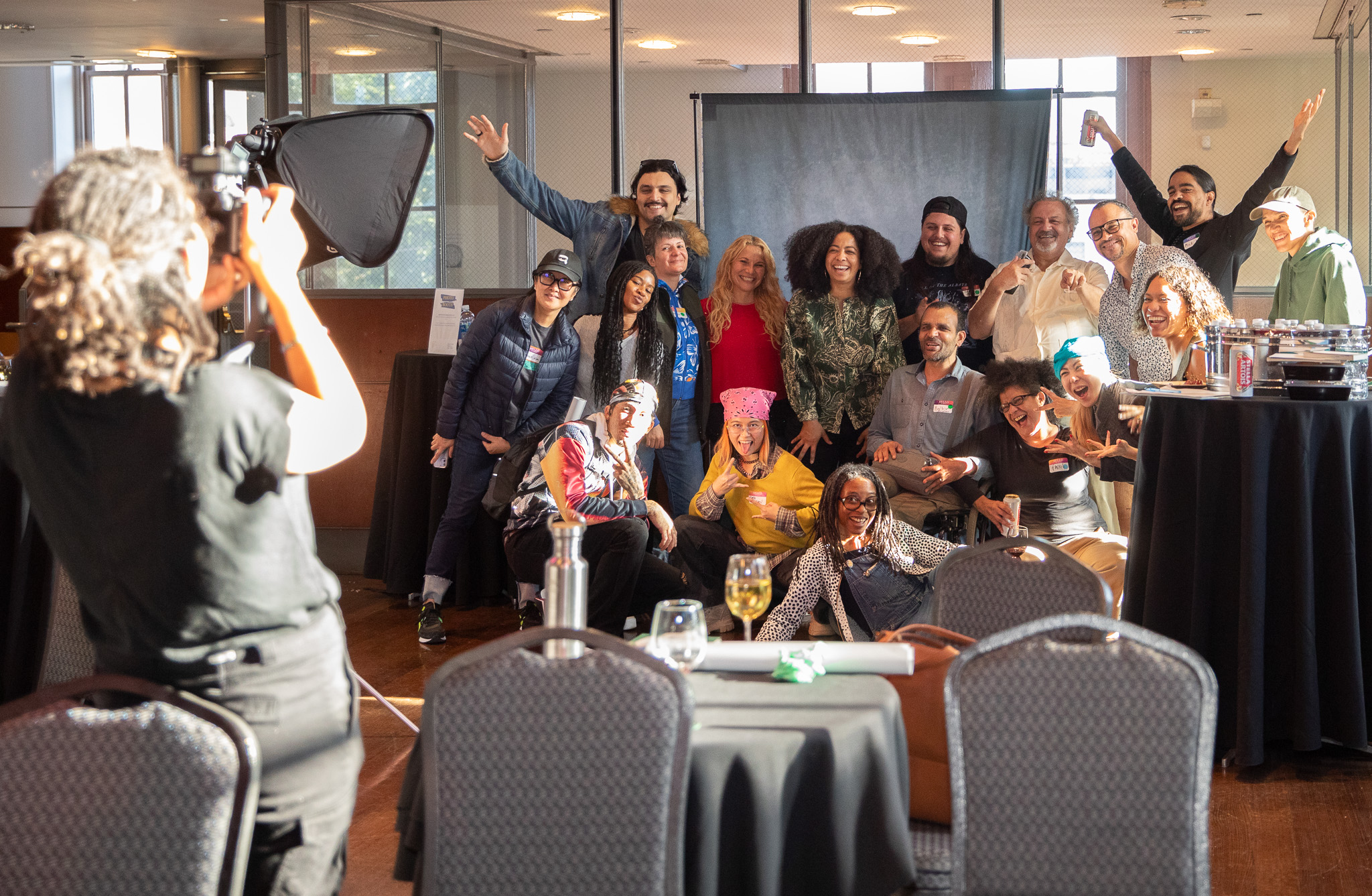 A professional photographer dressed in black stands in the foreground of the picture, photographing a group of 19, smiling people, some of whom are laughing and...