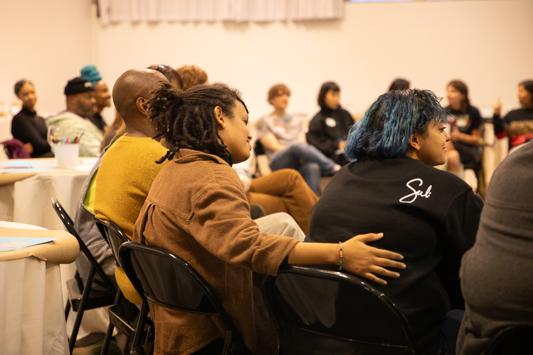 A group of people, facing away from the camera, are sitting in a circle; one person with locs, wearing a brown shirt, lovingly touches the back of another person...