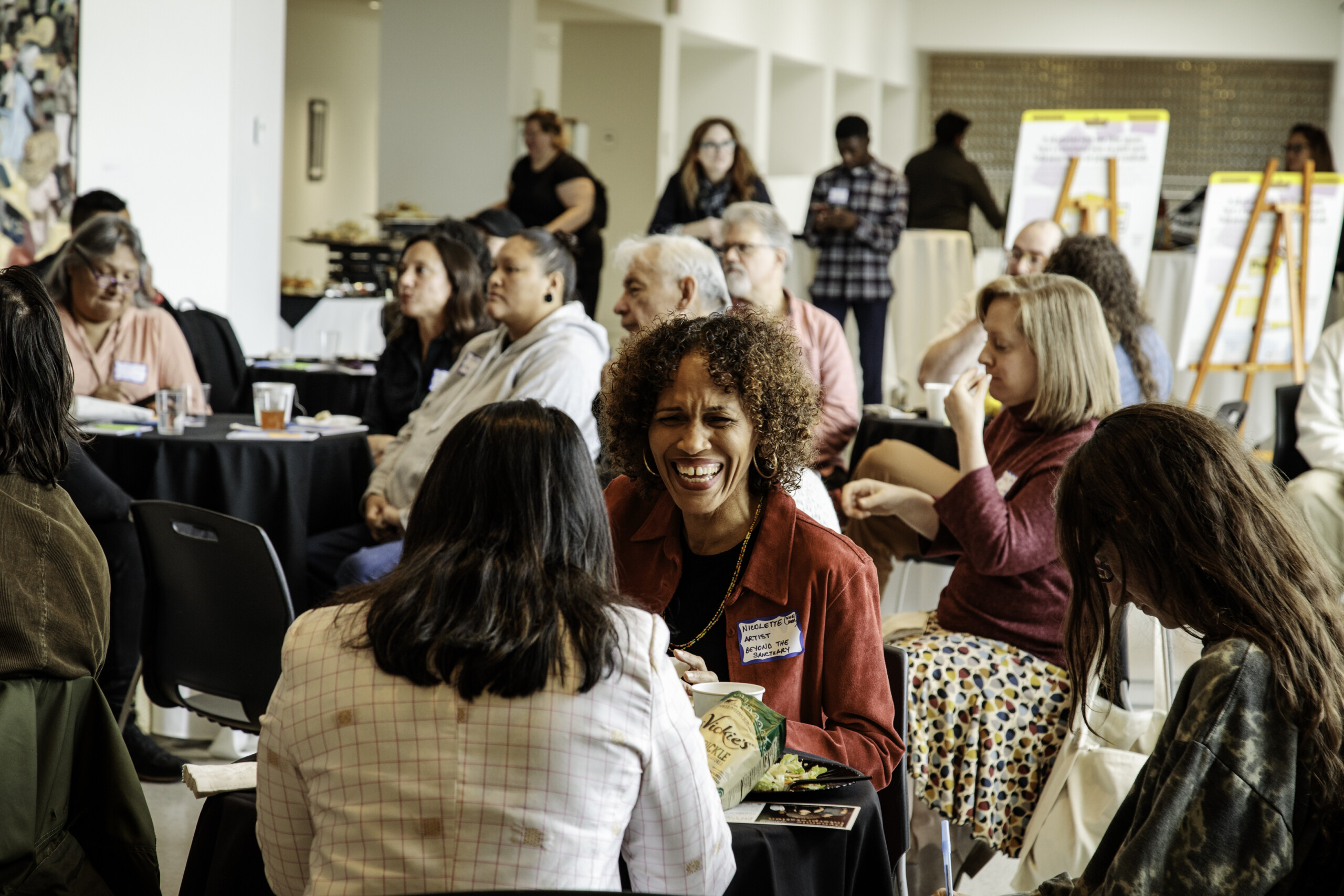 A smiling person with brown skin and natural hair dressed in a red, button-down top, a gold necklace, and gold hoop earrings talks to another person whose back...