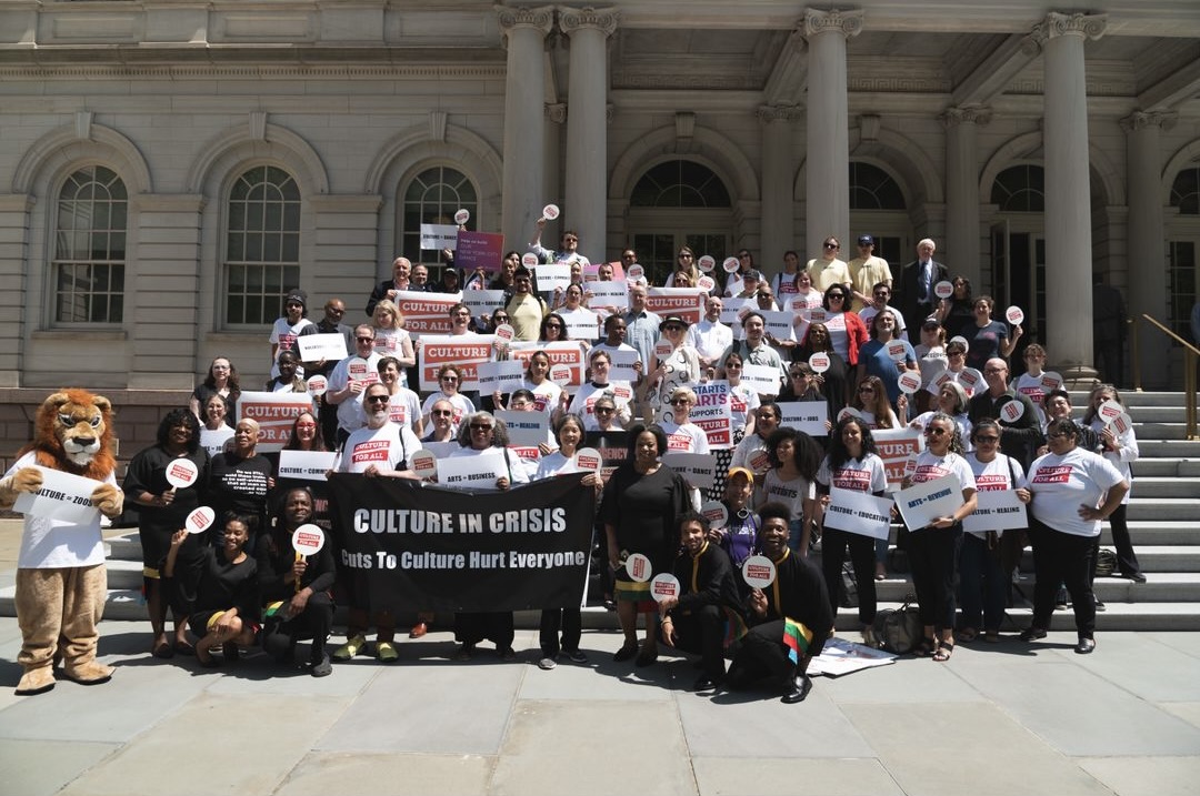 Photograph of a group of people wearing matching white shirts and holding signs with red text on a sunny day on the steps of City Hall. A lion mascot is seen on...