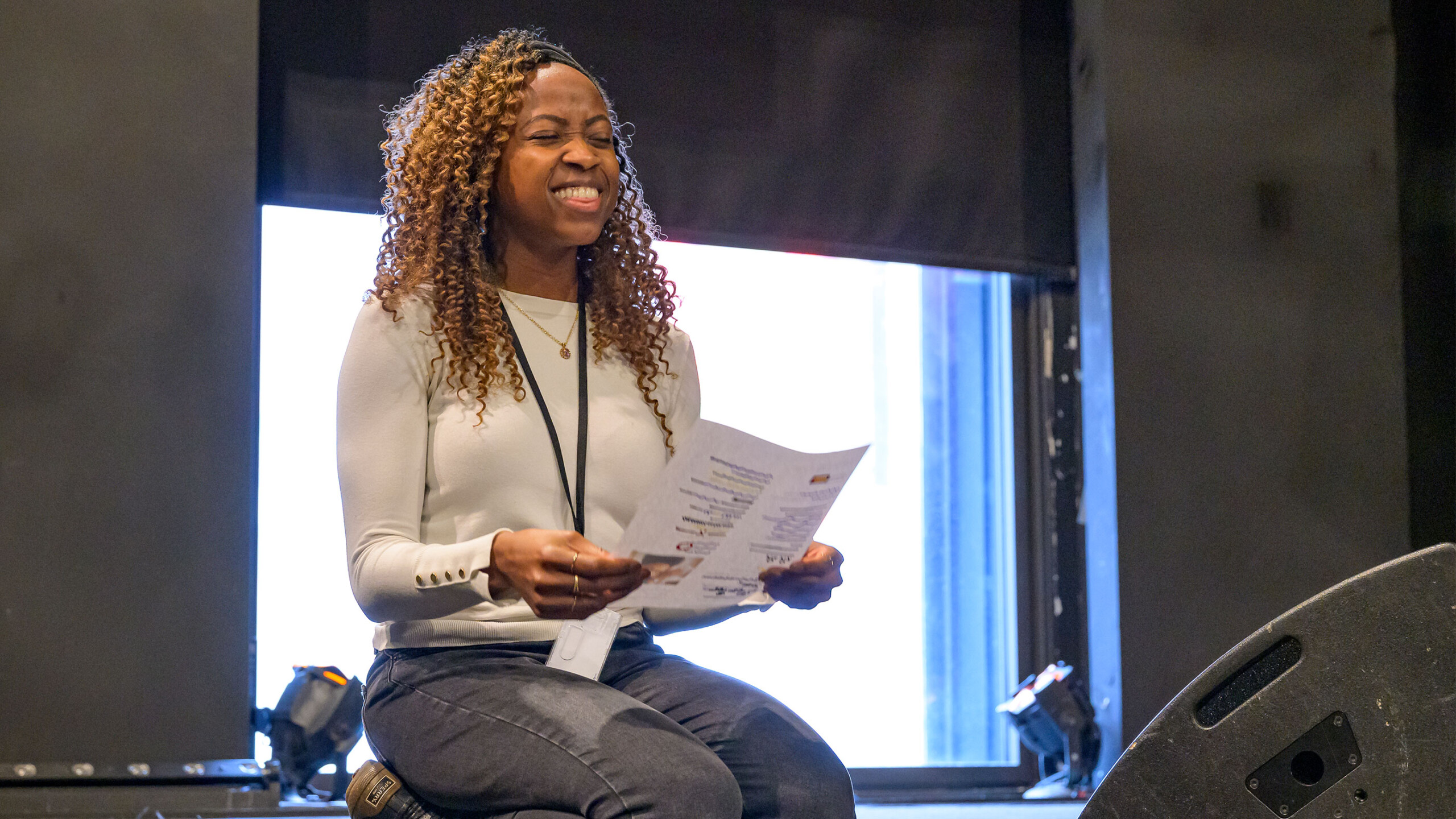 Woman sitting and reading a document indoors, next to a window and audio equipment.