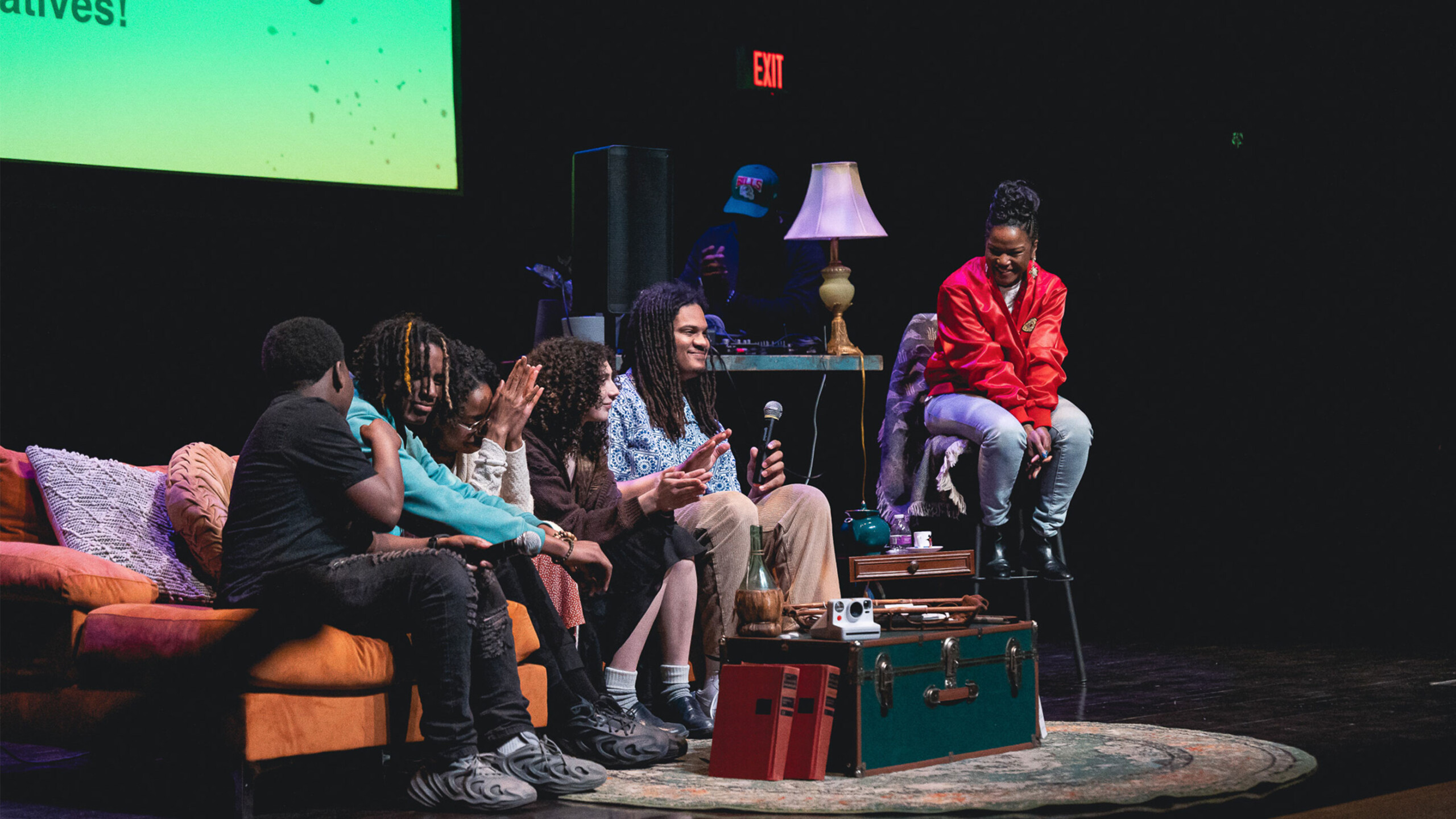 Group of people on stage with a couch, engaged in a discussion with a screen in the background.