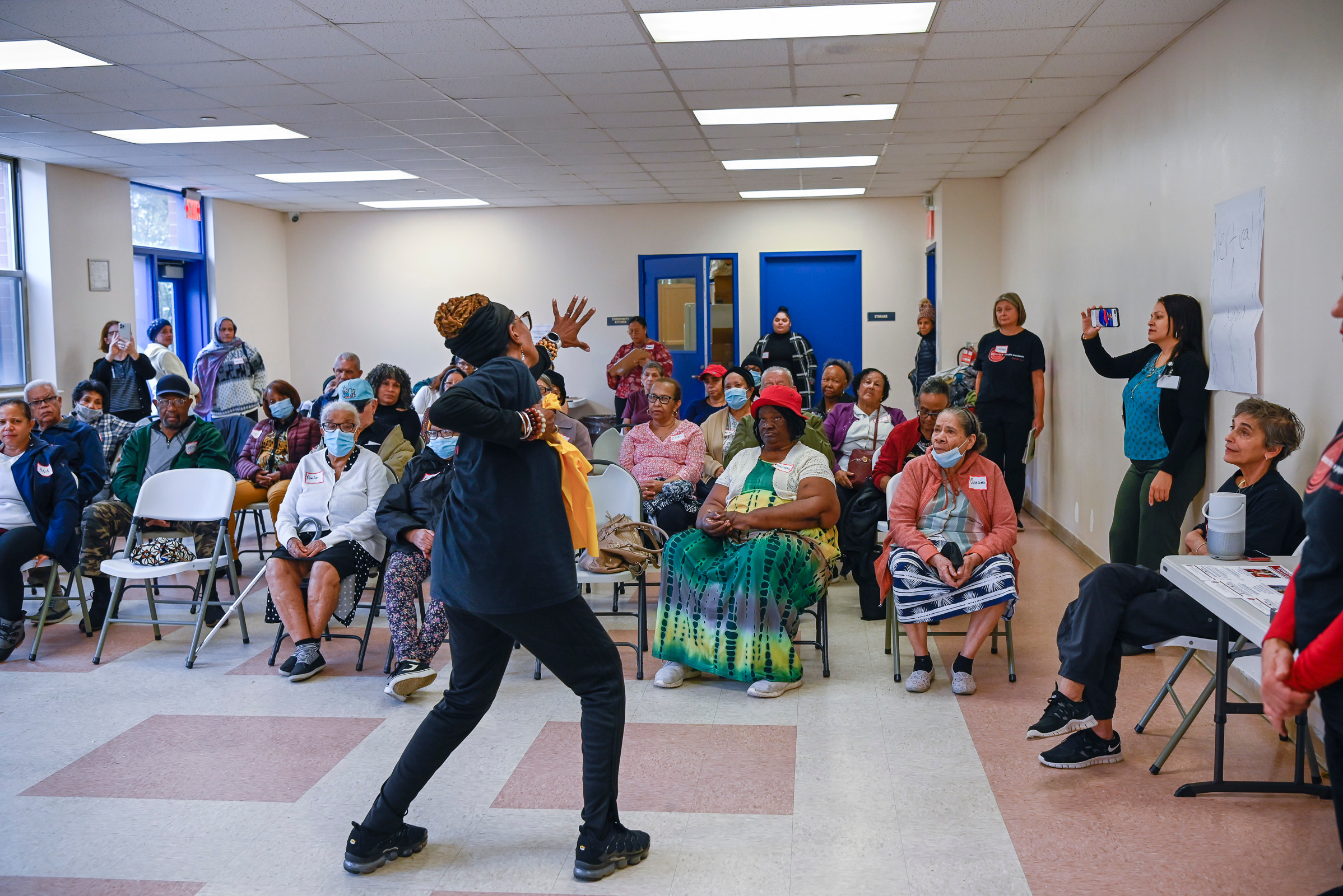 Indoor gathering with people seated in chairs watching a person gesturing energetically in the center.