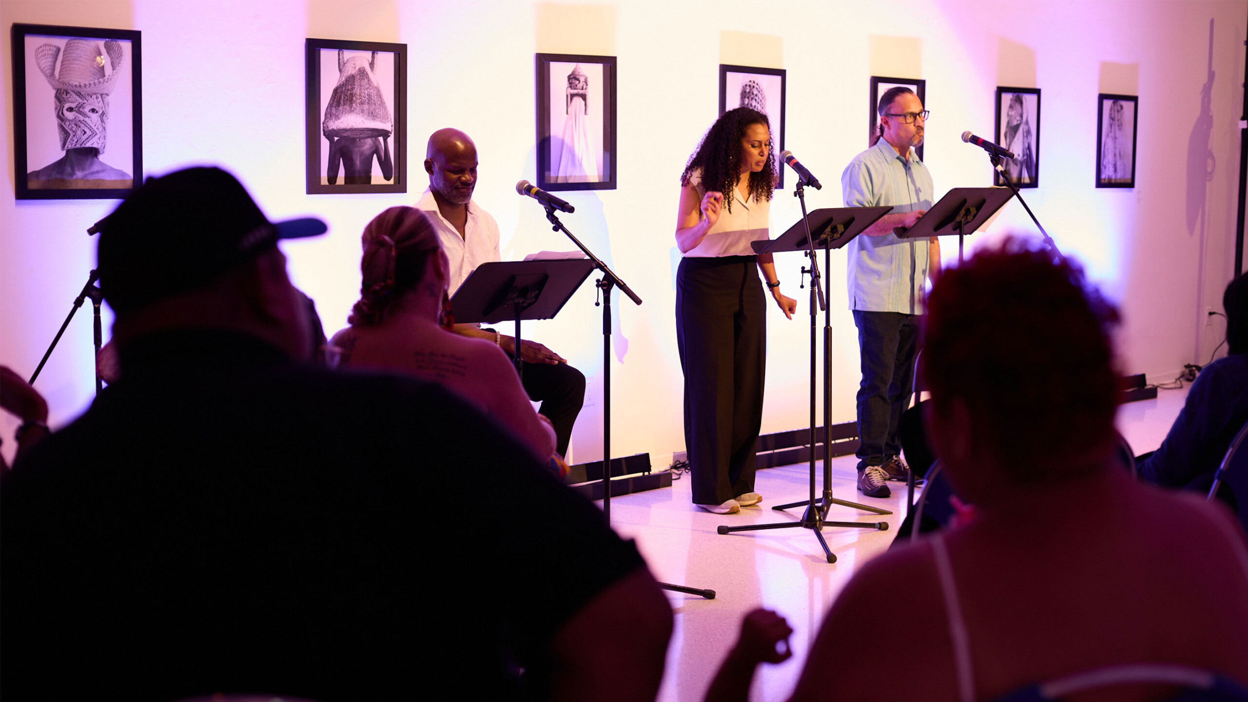 Three people on stage reading scripts by microphones; audience in foreground, art on walls.