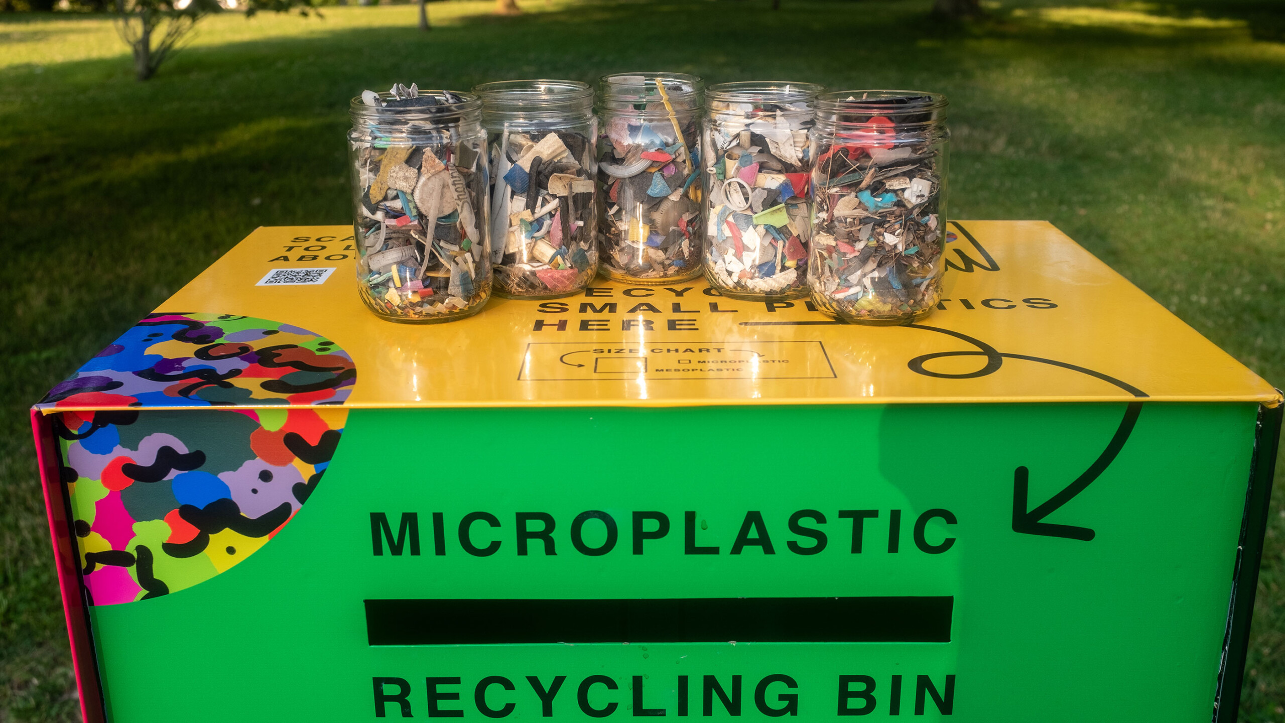 Five jars filled with microplastic pieces atop a colorful recycling bin.