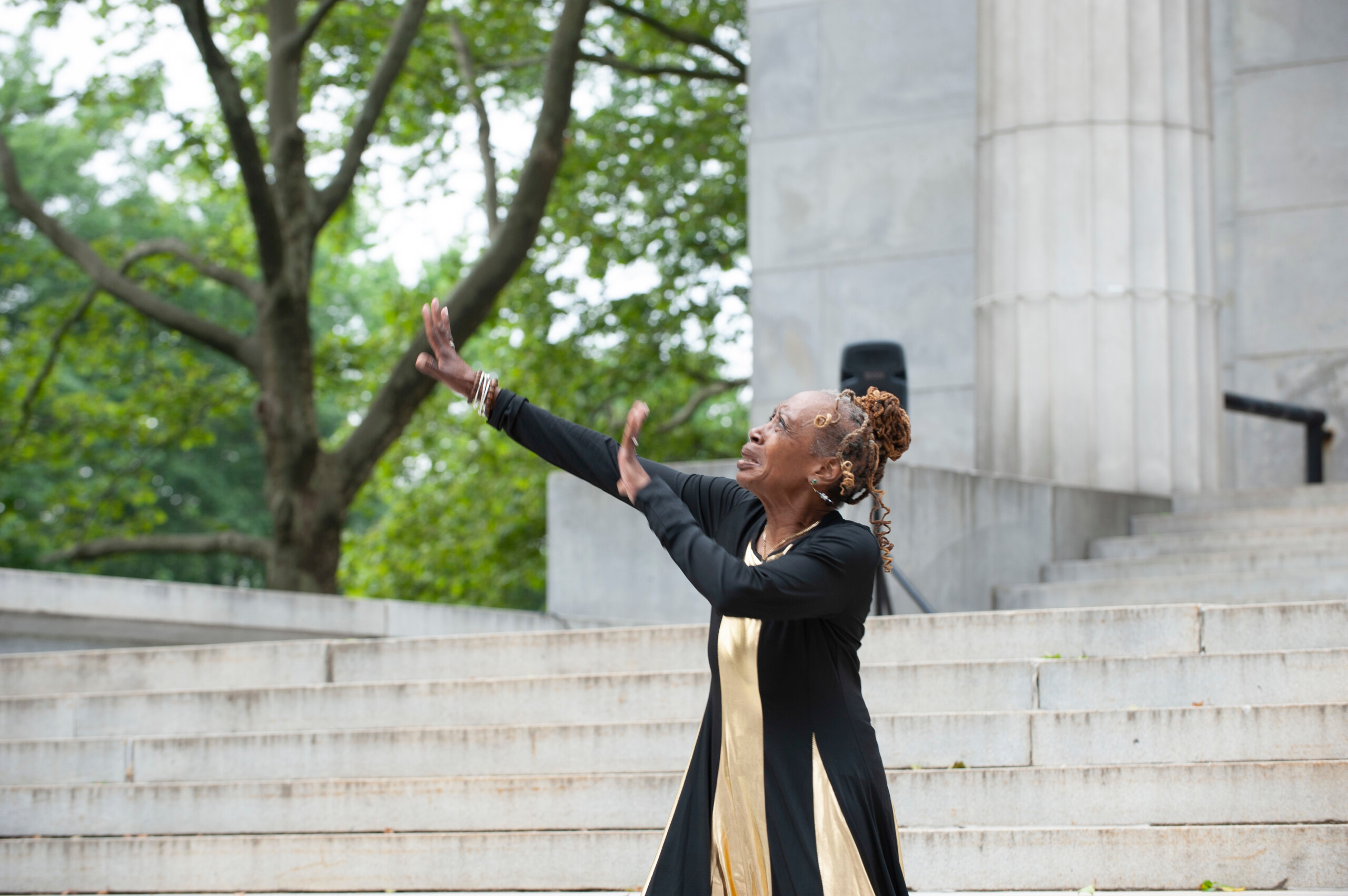Person in a black and gold dress performs a dance with arms outstretched on outdoor steps.