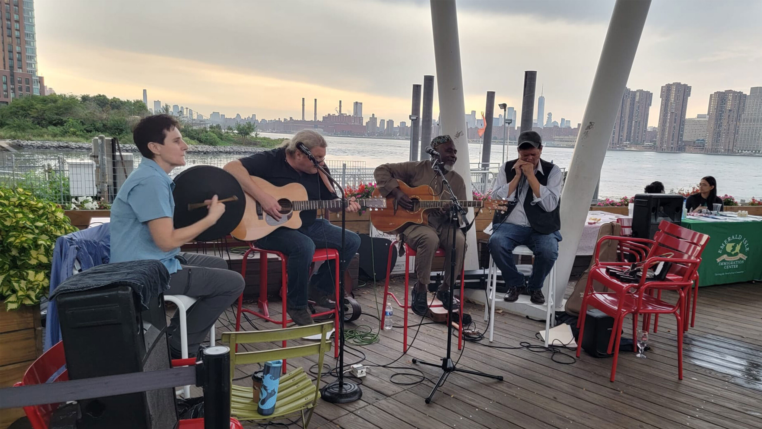 Musicians playing guitars on a riverside deck with city skyline in the background.
