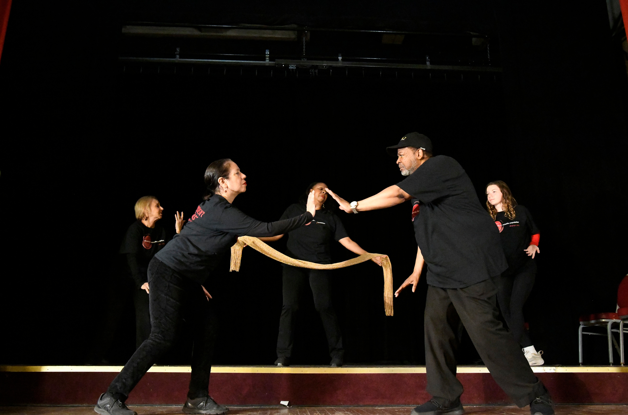 People on stage in black attire performing with a large wooden stick.