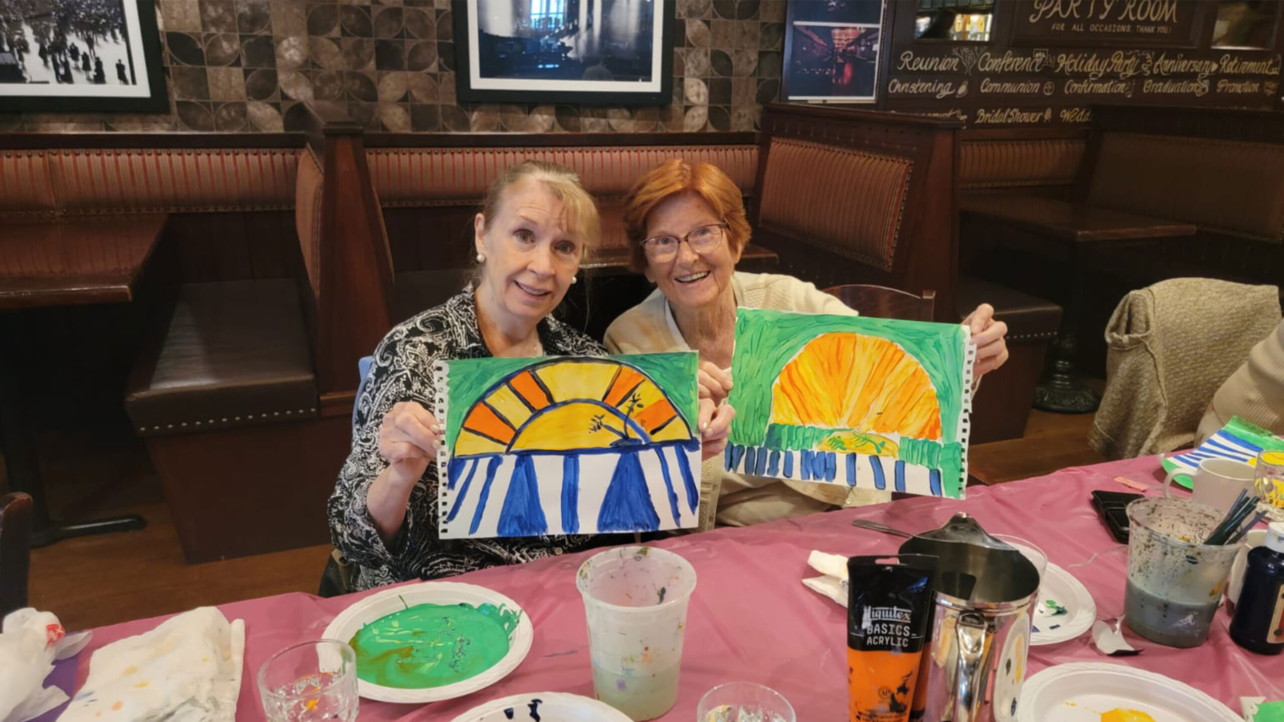 Two people holding up colorful paintings, surrounded by paint supplies on a table.