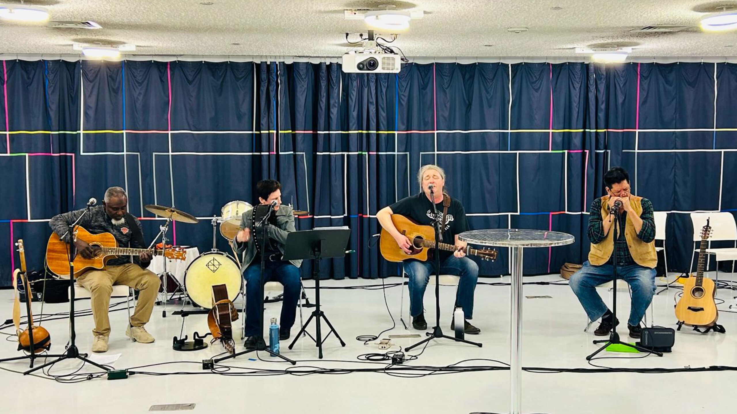Four musicians with guitars and a banjo in a room with blue curtains and a projector.