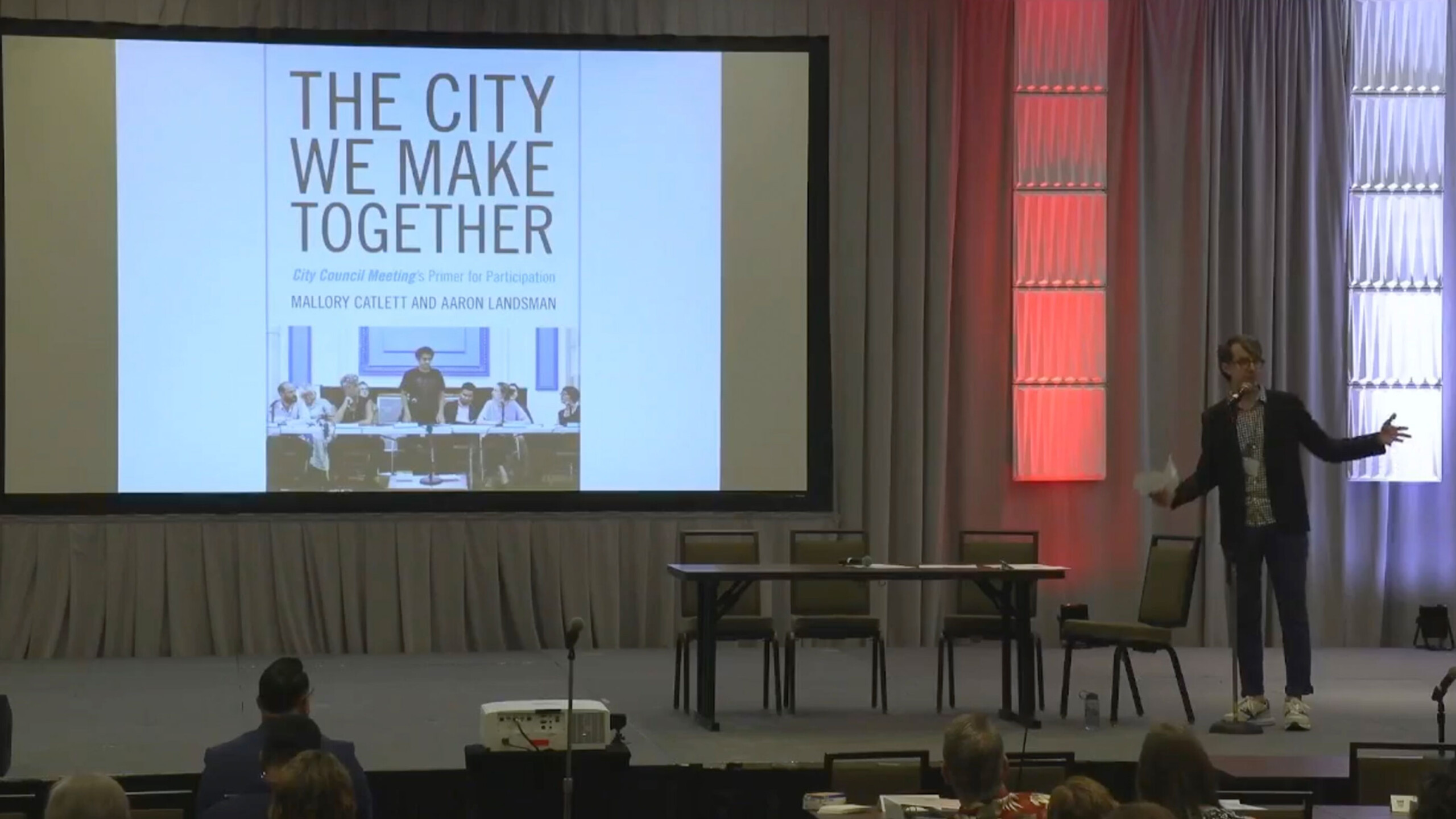 A speaker engages an audience at a conference or seminar, standing next to a large screen displaying a slide titled 'The City We Make Together' by Mallory Catlett...