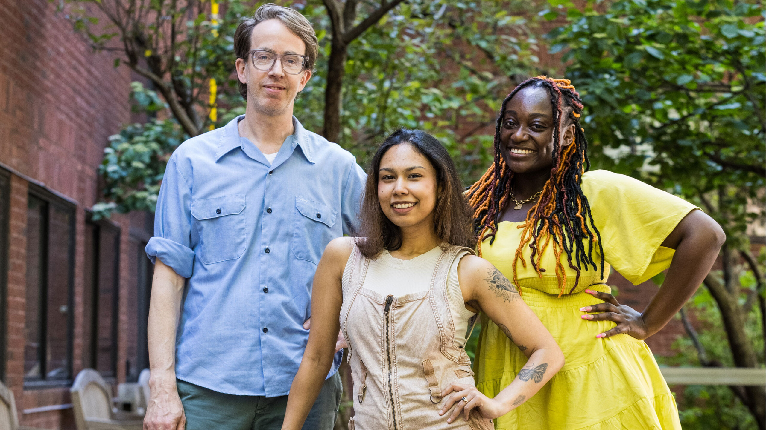 Three people are standing together outdoors, posing for a group photo. On the left, a man with glasses is wearing a light blue button-up shirt with rolled-up sleeves...