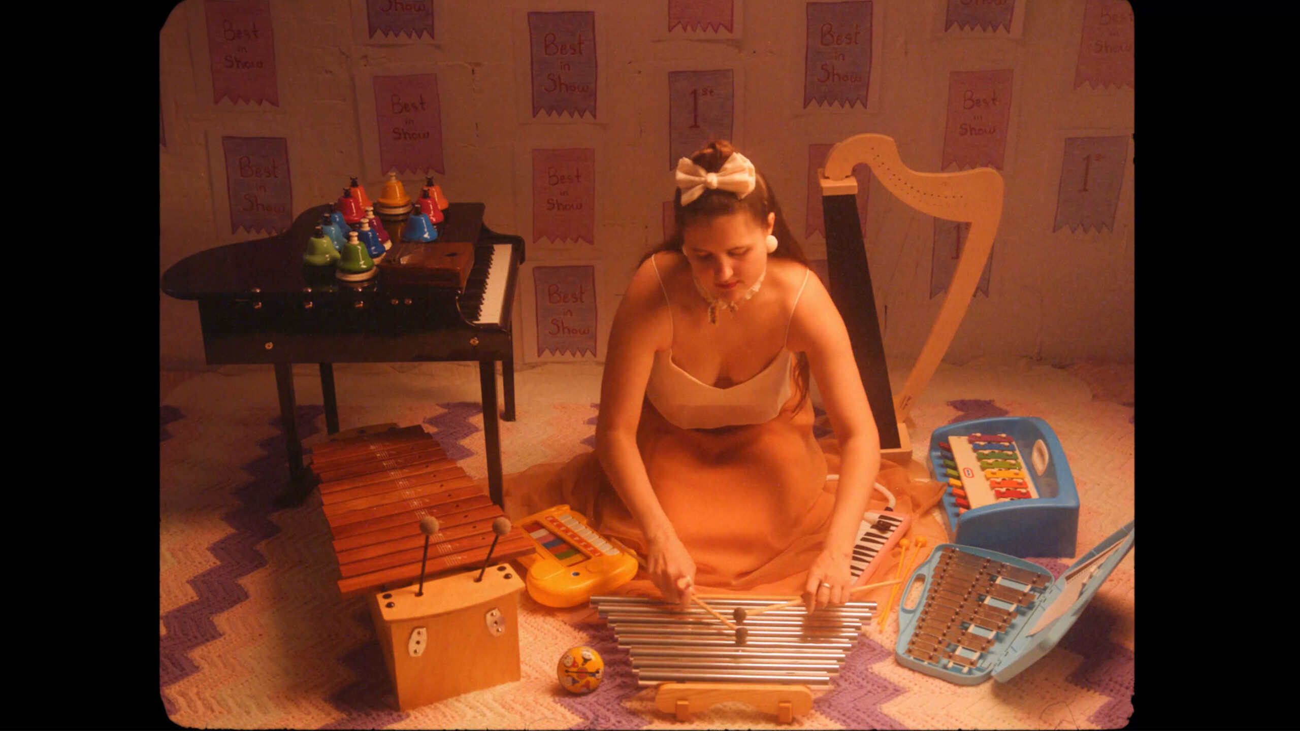 alexa dexa, in a silk gown with a white top and flowing peach skirtlength, kneels on the floor playing their toy chimes, Surrounding them from left to right are...