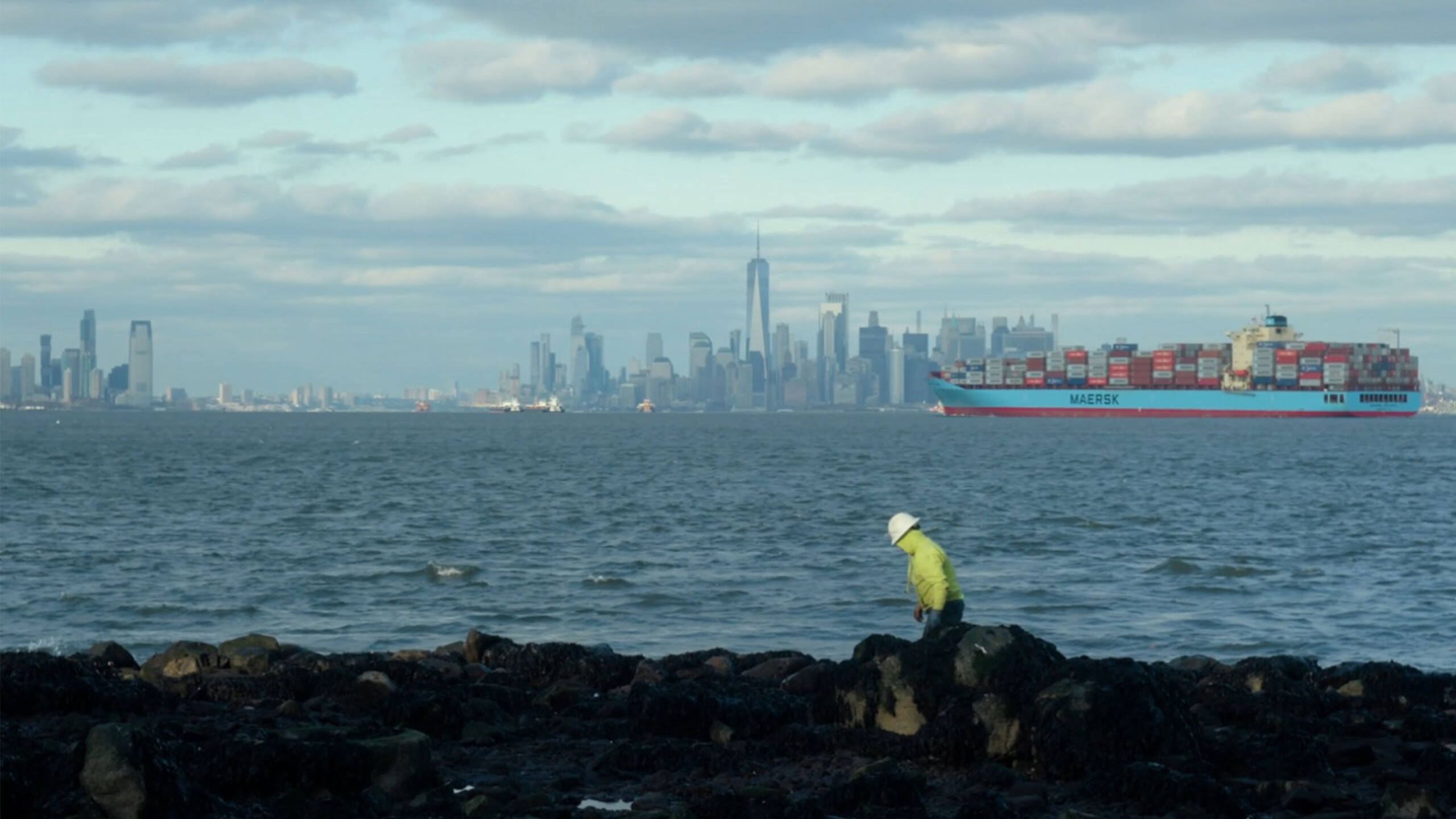 A person wearing a bright yellow jacket and white helmet is navigating over rocky terrain by the water's edge. In the background, a vast body of water separates...