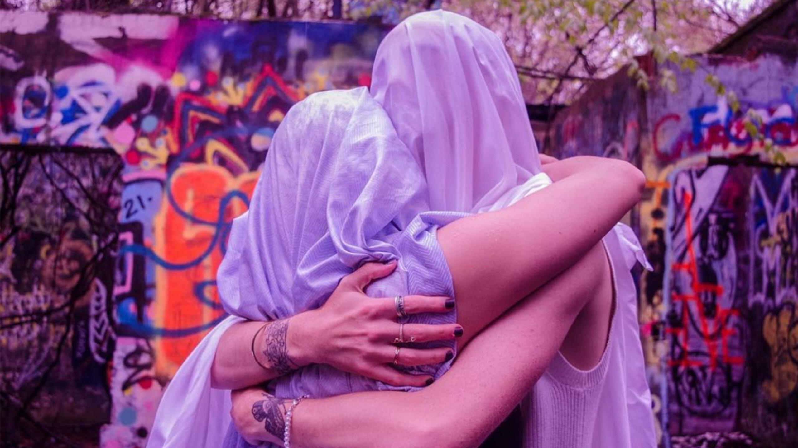 Two individuals wrapped in translucent white veils tightly embrace each other, creating a sense of unity and intimacy. Their arms are adorned with tattoos and rings,...