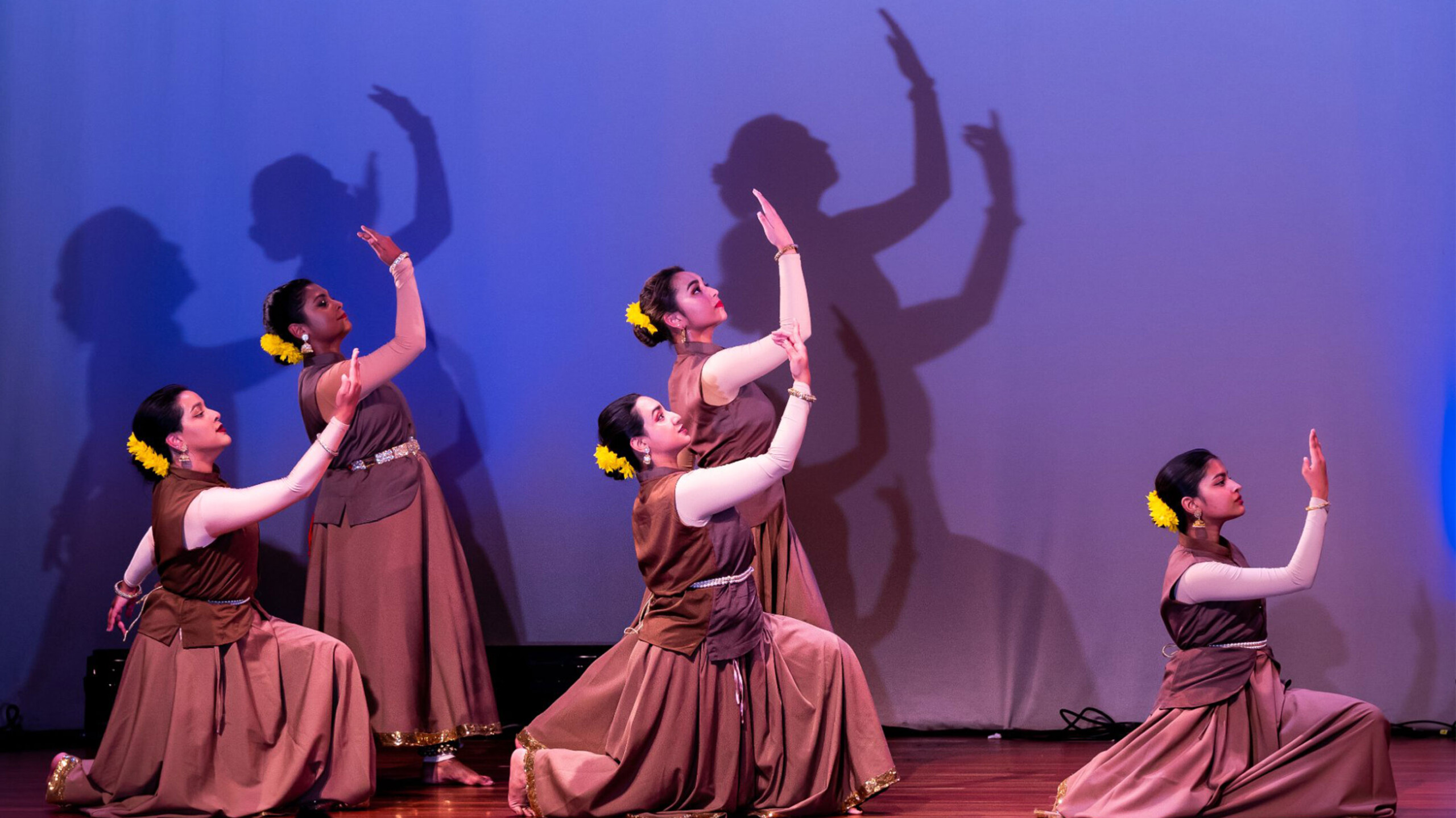 Dancers in brown outfits performing with shadows projected on a blue background.