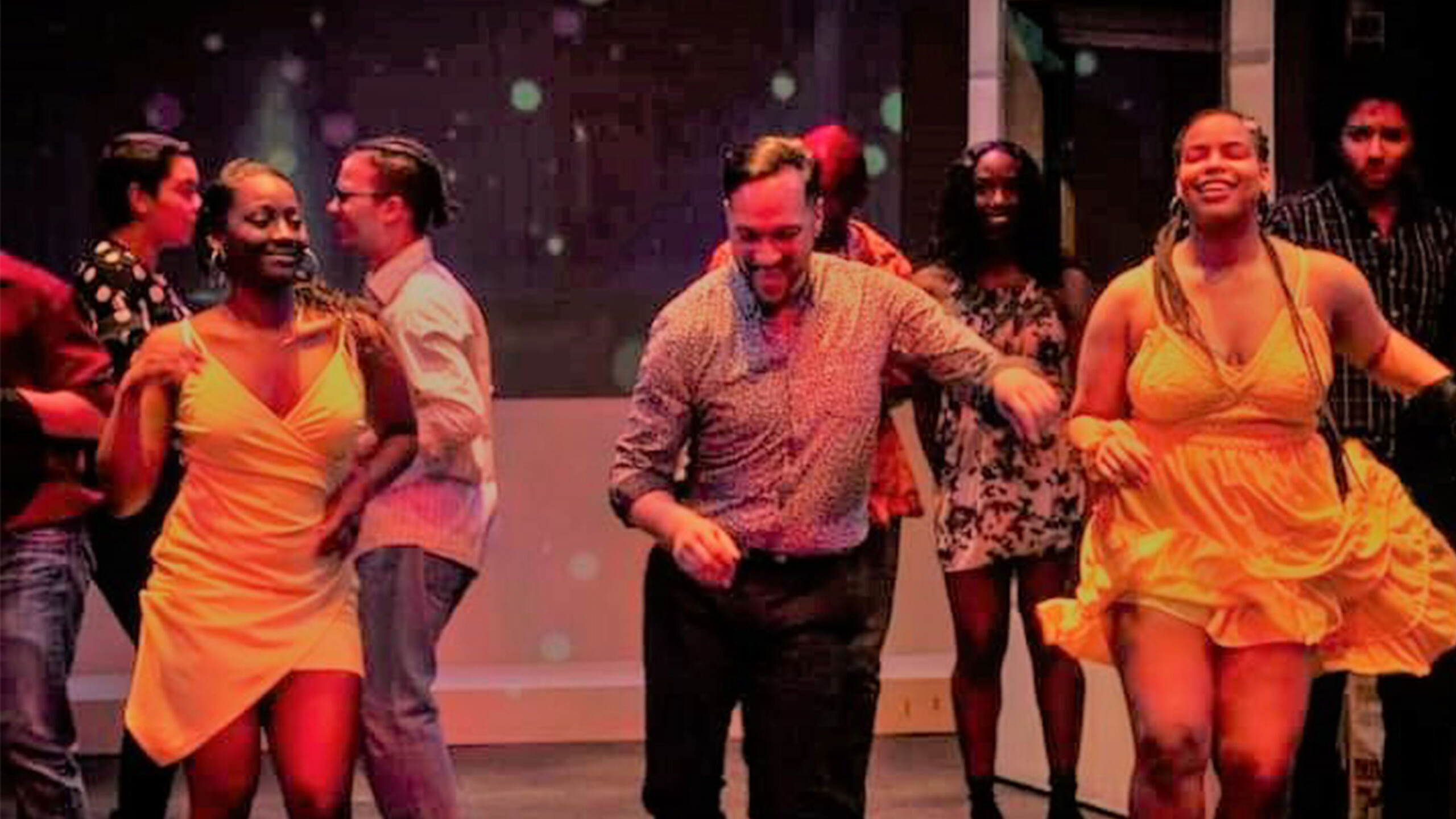 A close up of three dancers, caught mid motion. The two dancers flanking the central figure wear bright yellow dresses. All three dancers wear a joyful expression....