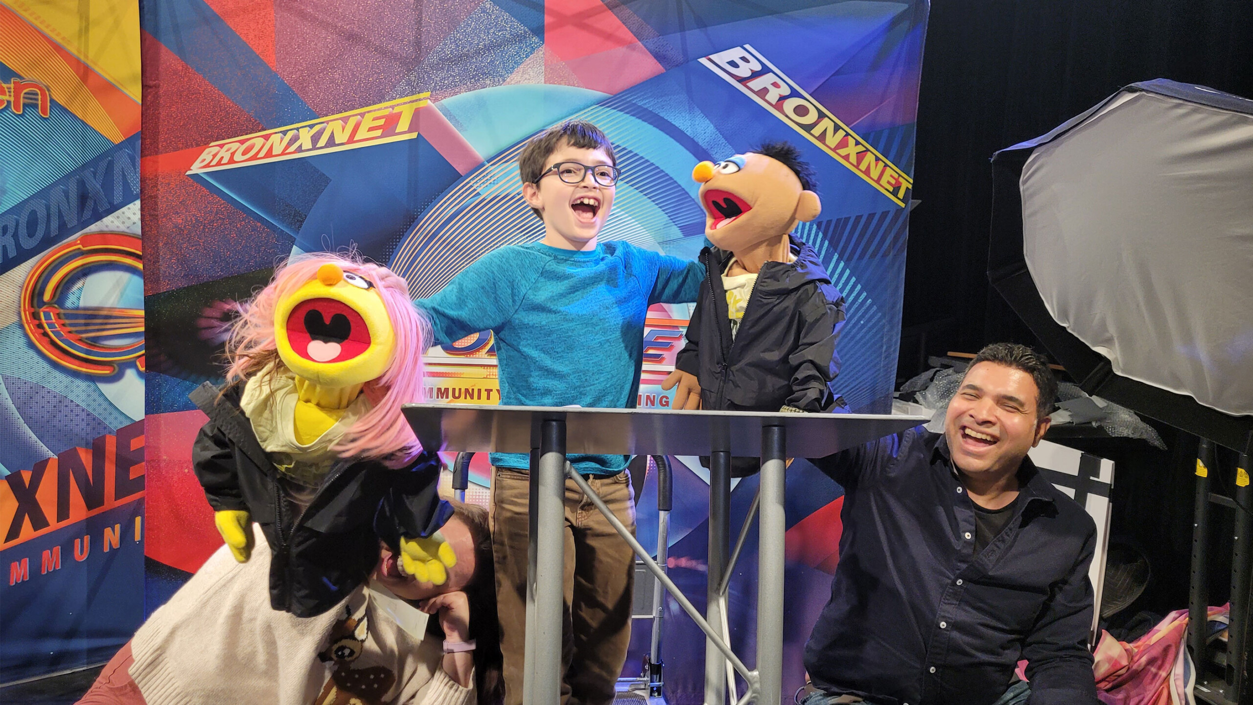A lively scene at a television studio features two cheerful puppets with human-like features entertaining a young boy wearing glasses and a teal shirt. The boy...