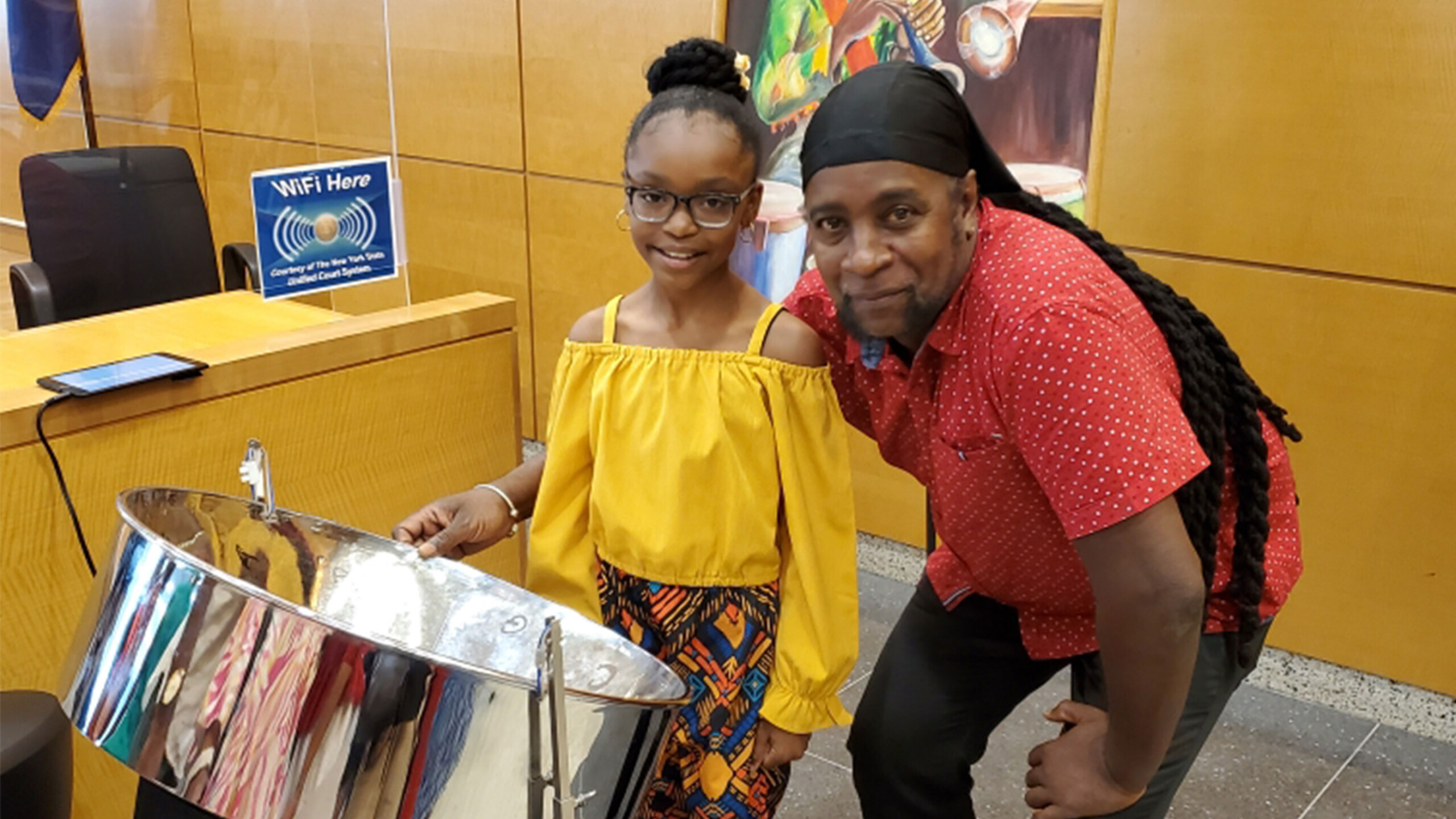 A young girl and an adult man are posing with a steel drum in an indoor setting. The girl is wearing a yellow off-shoulder top and patterned pants, while the man...