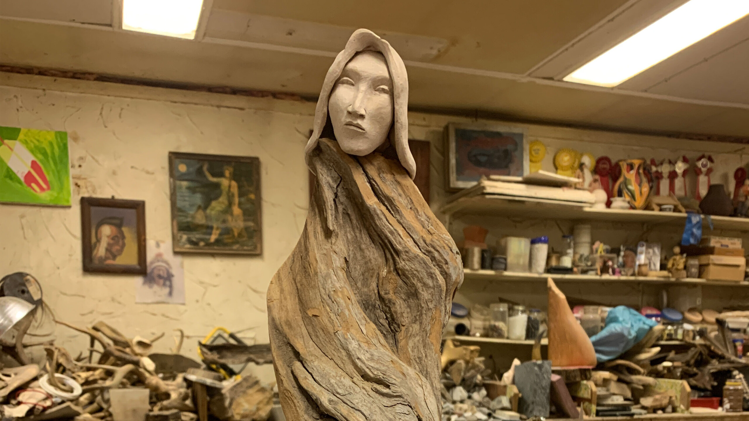 A striking sculpture stands prominently against the backdrop of a cluttered artist's workshop. The sculpture features a serene human face carved from smooth stone...