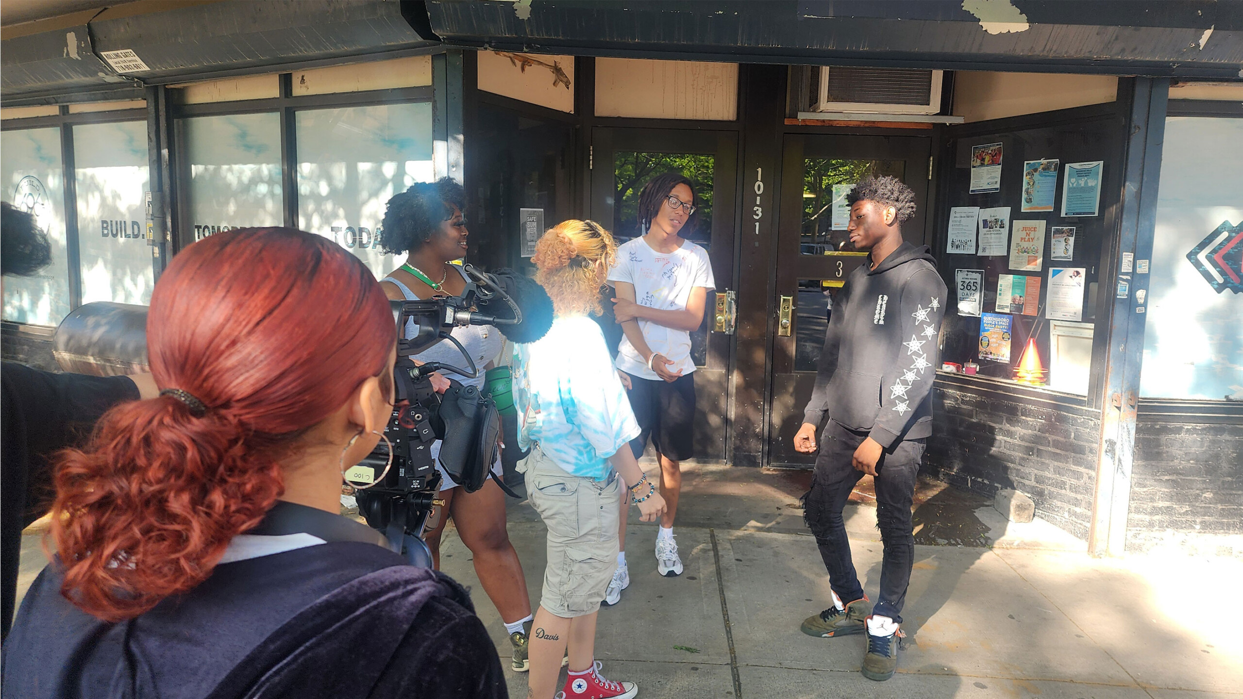 A small group of people is gathered outside a building, filming a scene. A person with a camera films two individuals in conversation in the center, while another...