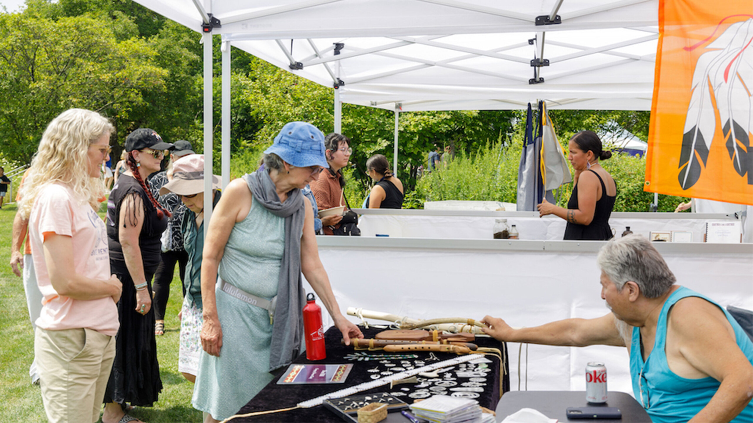 An outdoor market stall features a variety of handmade goods displayed on a table, including small crafts and what appears to be wooden or bone items. A man seated...