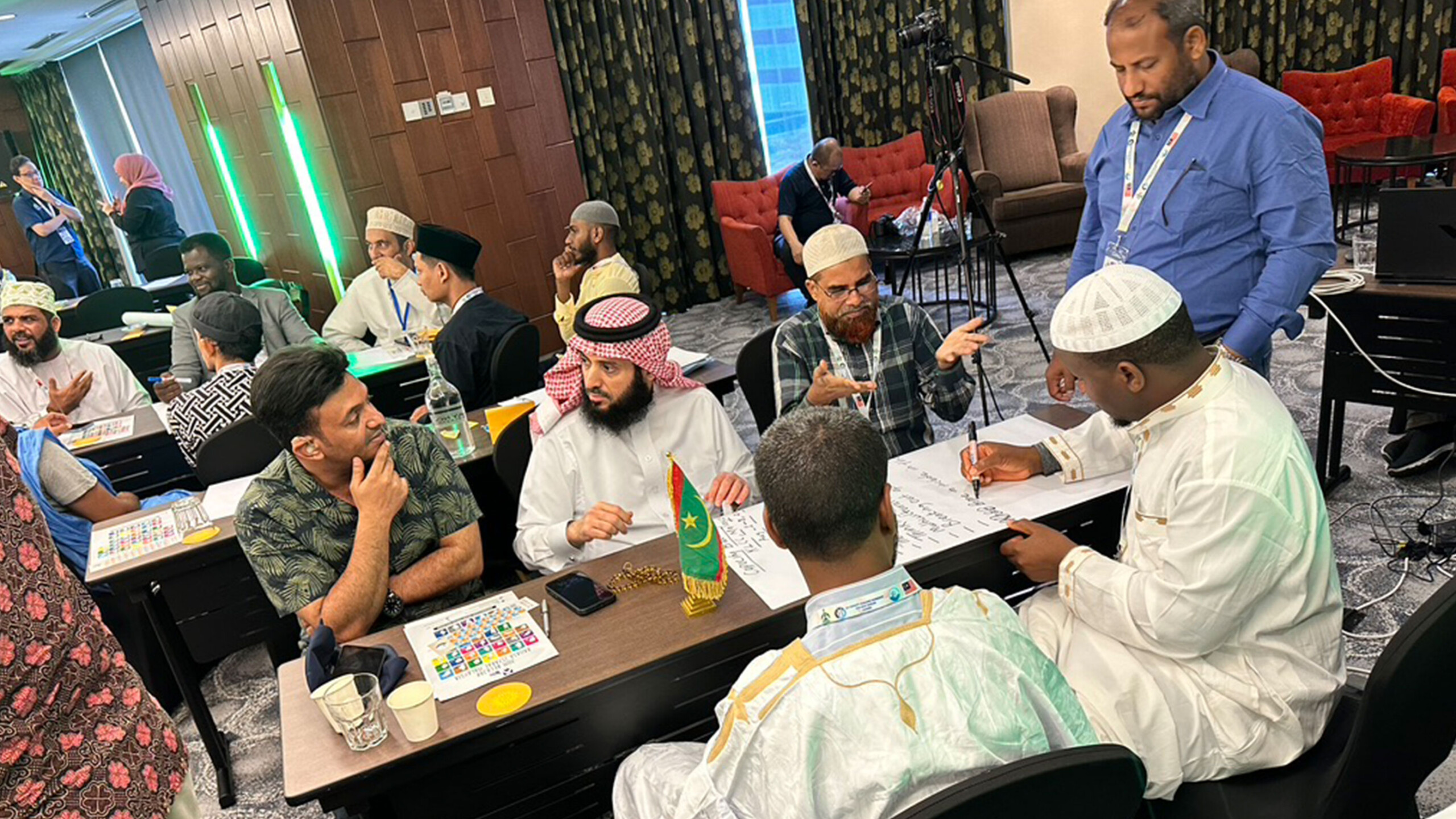 A diverse group of individuals is engaged in conversation around tables in an indoor setting, possibly a conference or workshop. Participants are of various cultural...