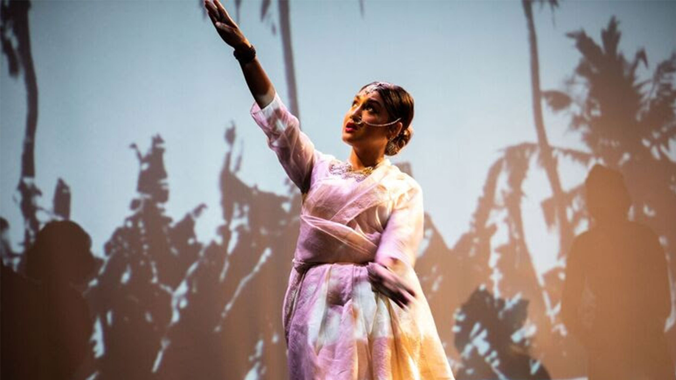 A woman stands in front of a projected image of black and white palm trees. She is dressed in a light pink sari. She stands in the center of the image, eyes gazing...