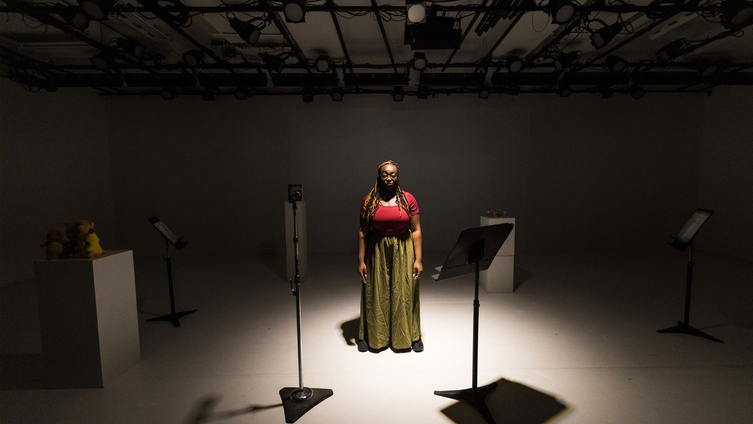 A lone performer stands in the center of a minimally lit studio. Bright spotlights overhead illuminate her, casting distinct shadows behind. She wears a flowing...