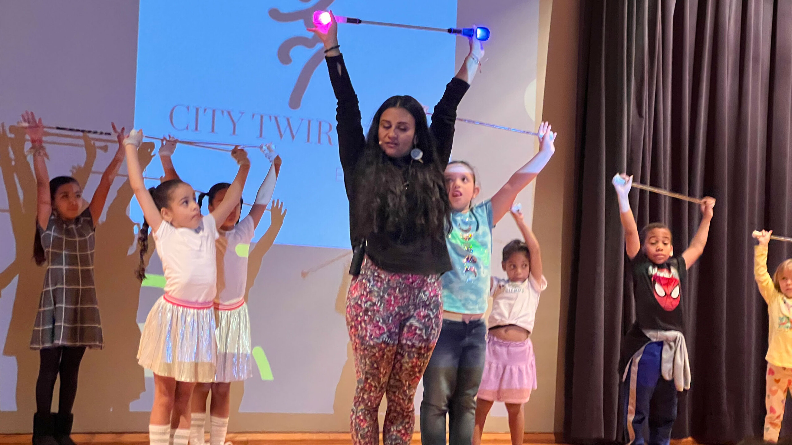 A group of young children and their instructor on stage, participating in a baton twirling performance. The instructor stands at the center with a baton held high...