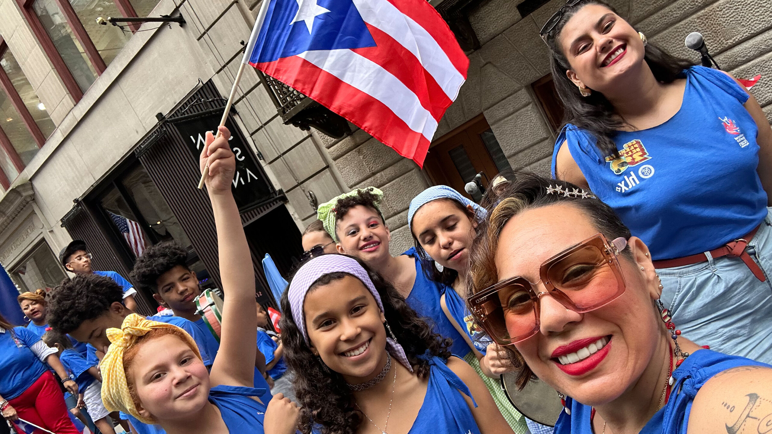 A group of smiling individuals, wearing matching blue shirts, gather on a city street, participating in a festive event. One woman in the forefront wears sunglasses...