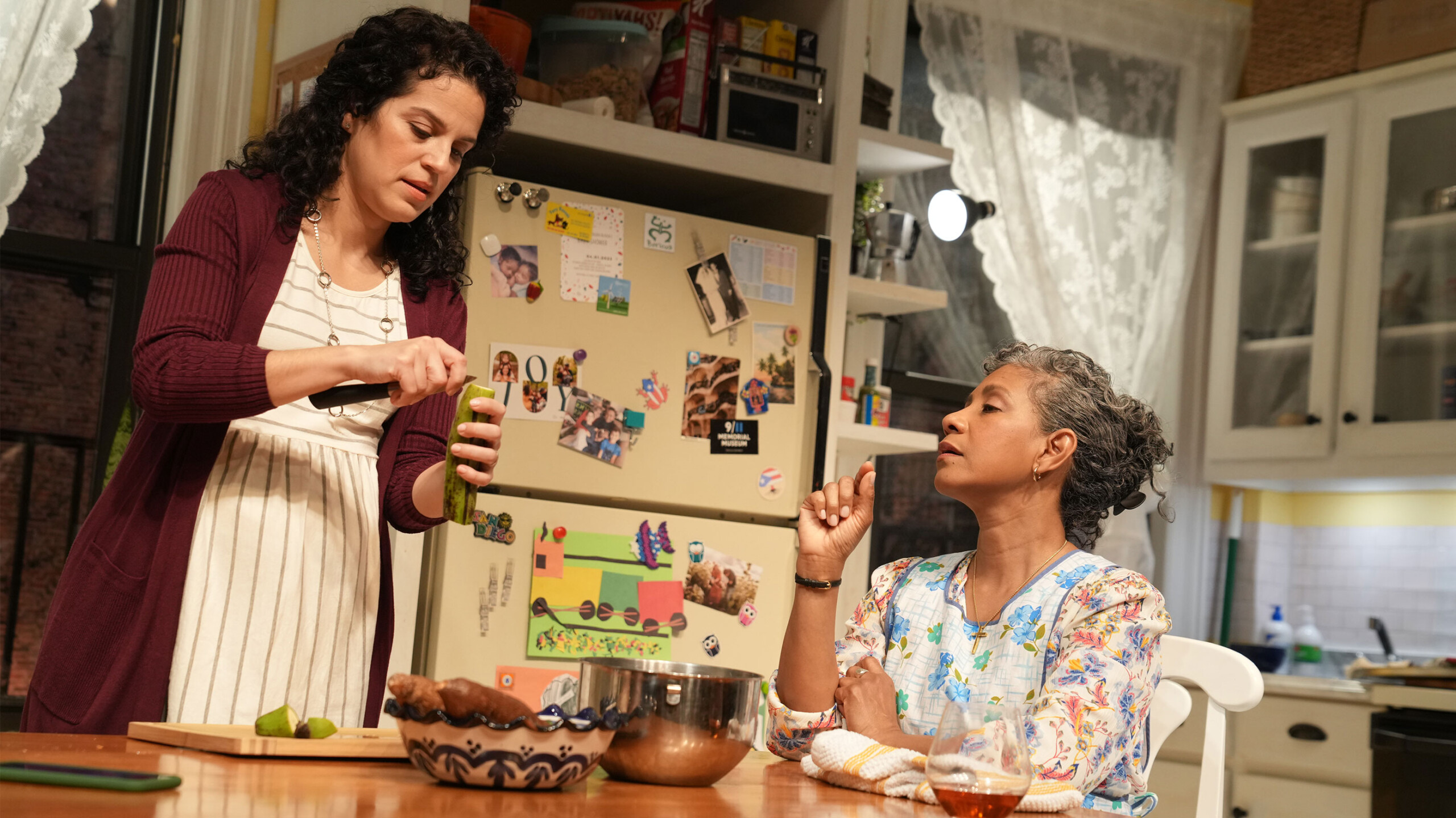 Two individuals in a kitchen with one cutting limes and a refrigerator adorned with photos and magnets.