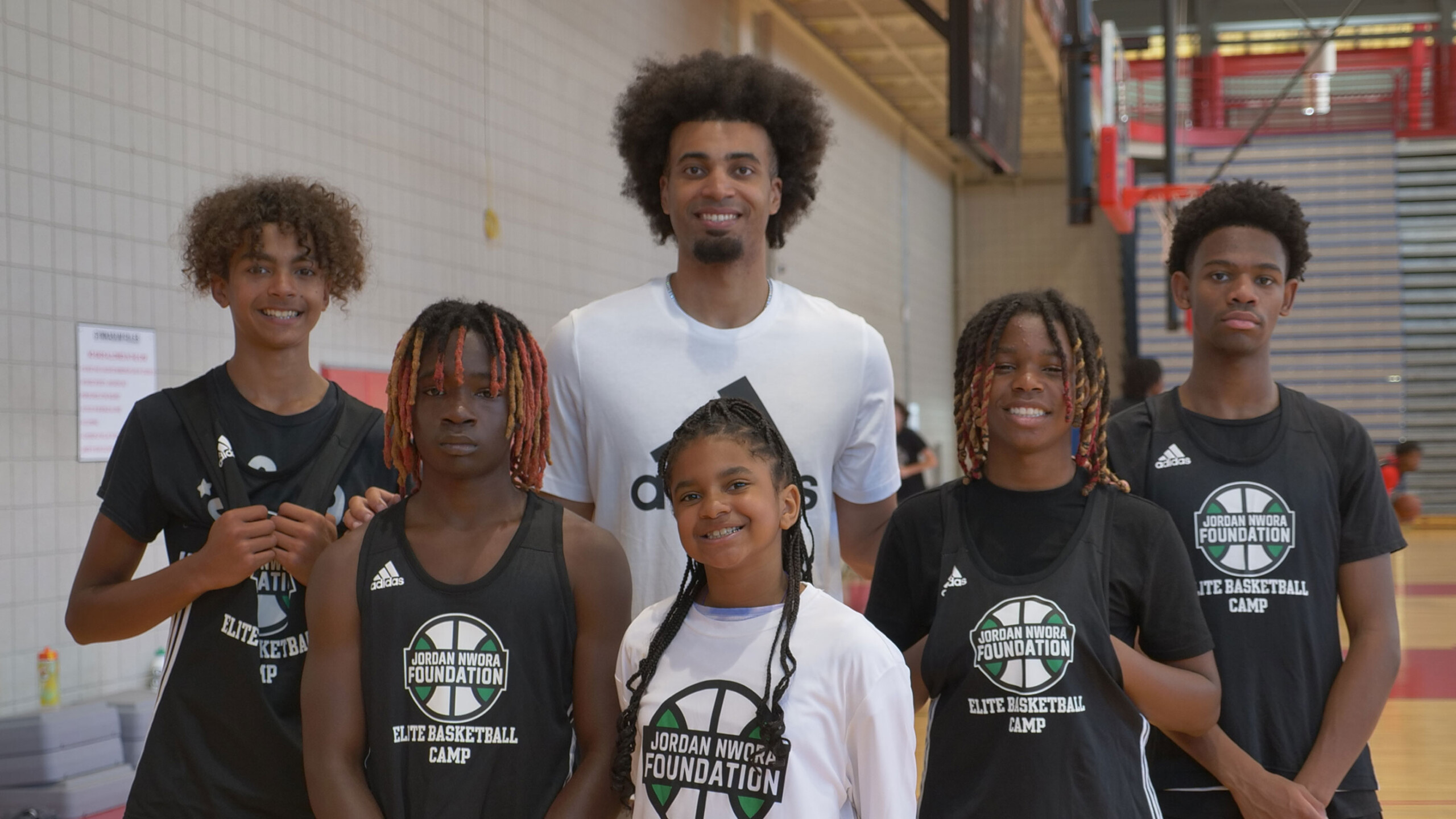 A group of young basketball players and a coach stand together in a gymnasium, forming a cheerful group for a photo. The players, five in total, are wearing matching...
