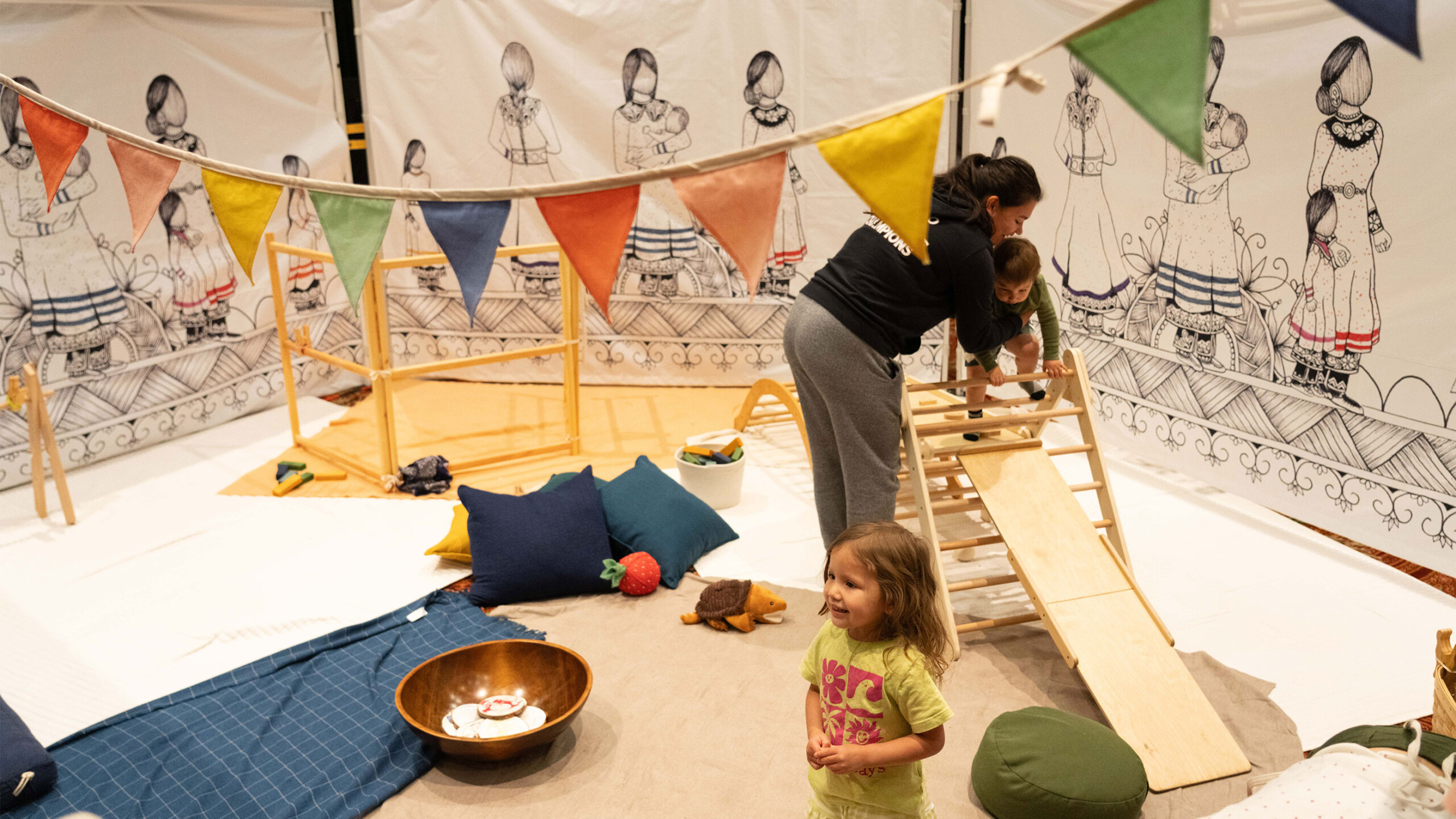 A cozy indoor play area features a woman helping a young child climb a wooden slide. Another child stands nearby, wearing a bright T-shirt. The space is decorated...