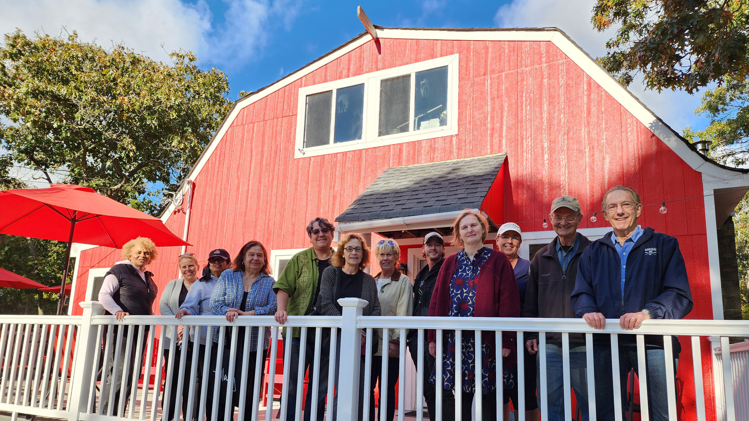 Group of people gathered on a white-fenced deck in front of a red barn under a sunny sky.