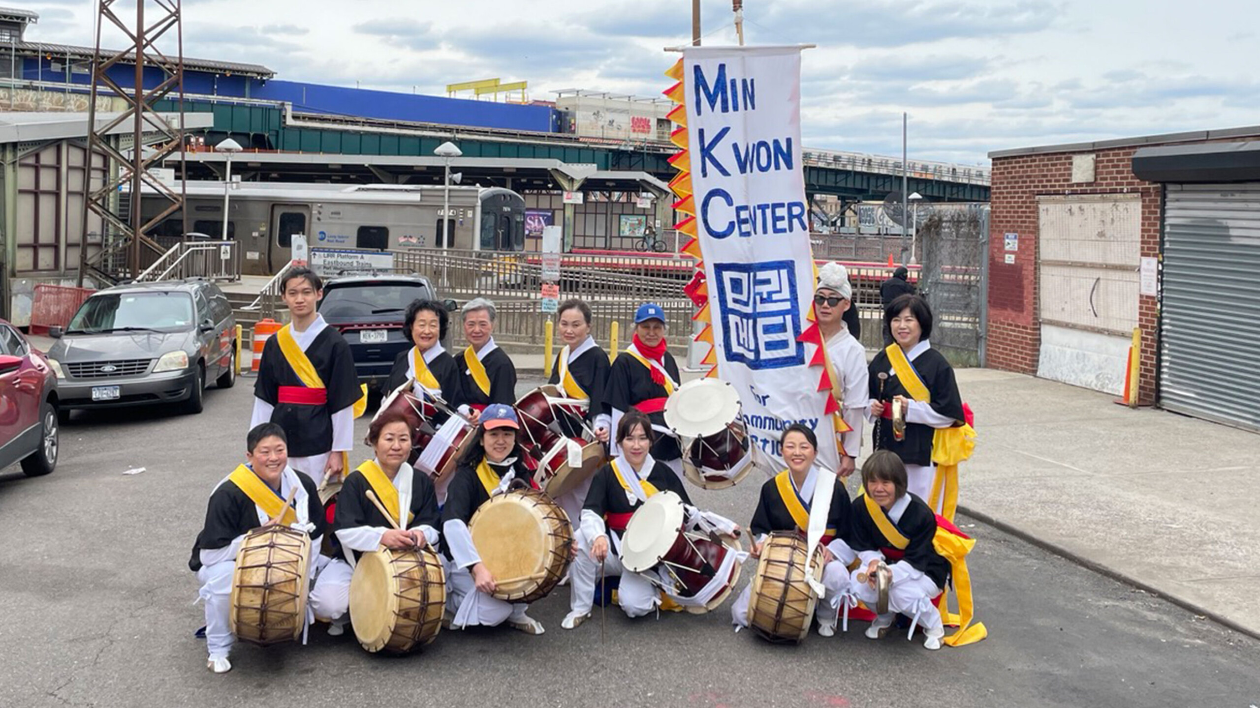 A group of people dressed in traditional Korean percussion attire, featuring black, white, and yellow costumes with red accents. Many hold traditional Korean drums...