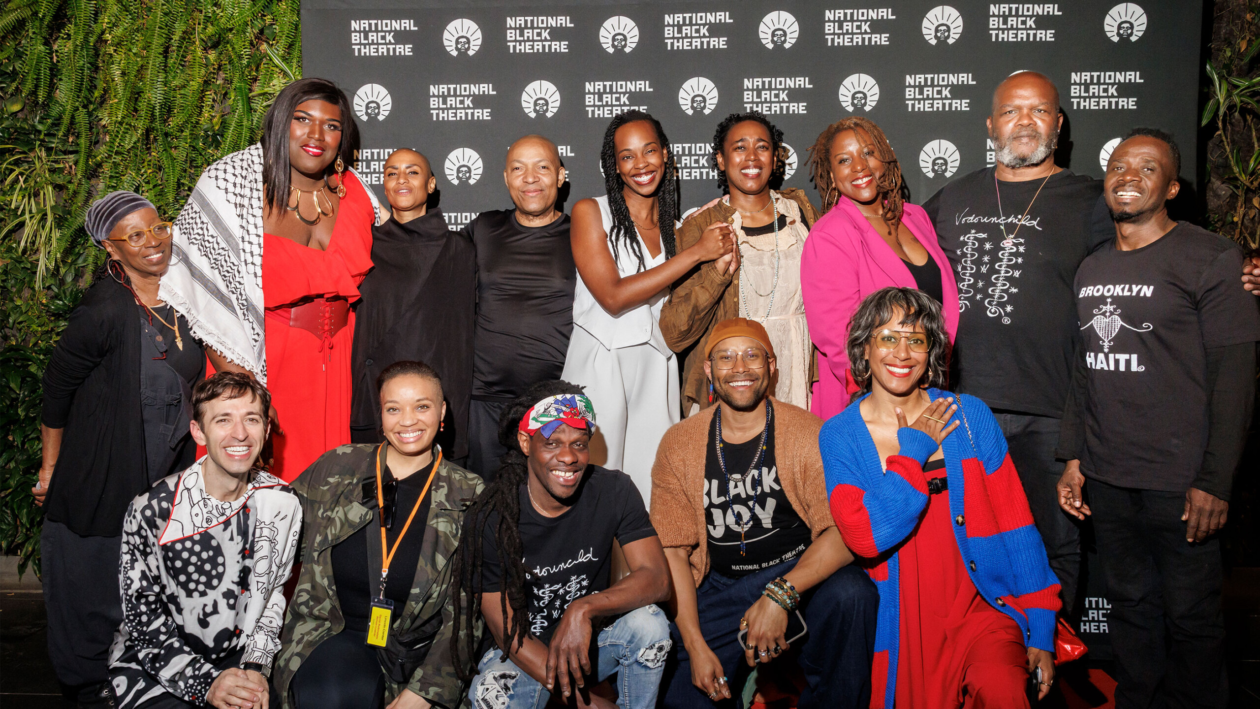 A diverse group of individuals poses together in front of a backdrop featuring the National Black Theatre logo. The people in the photo are wearing a variety of...