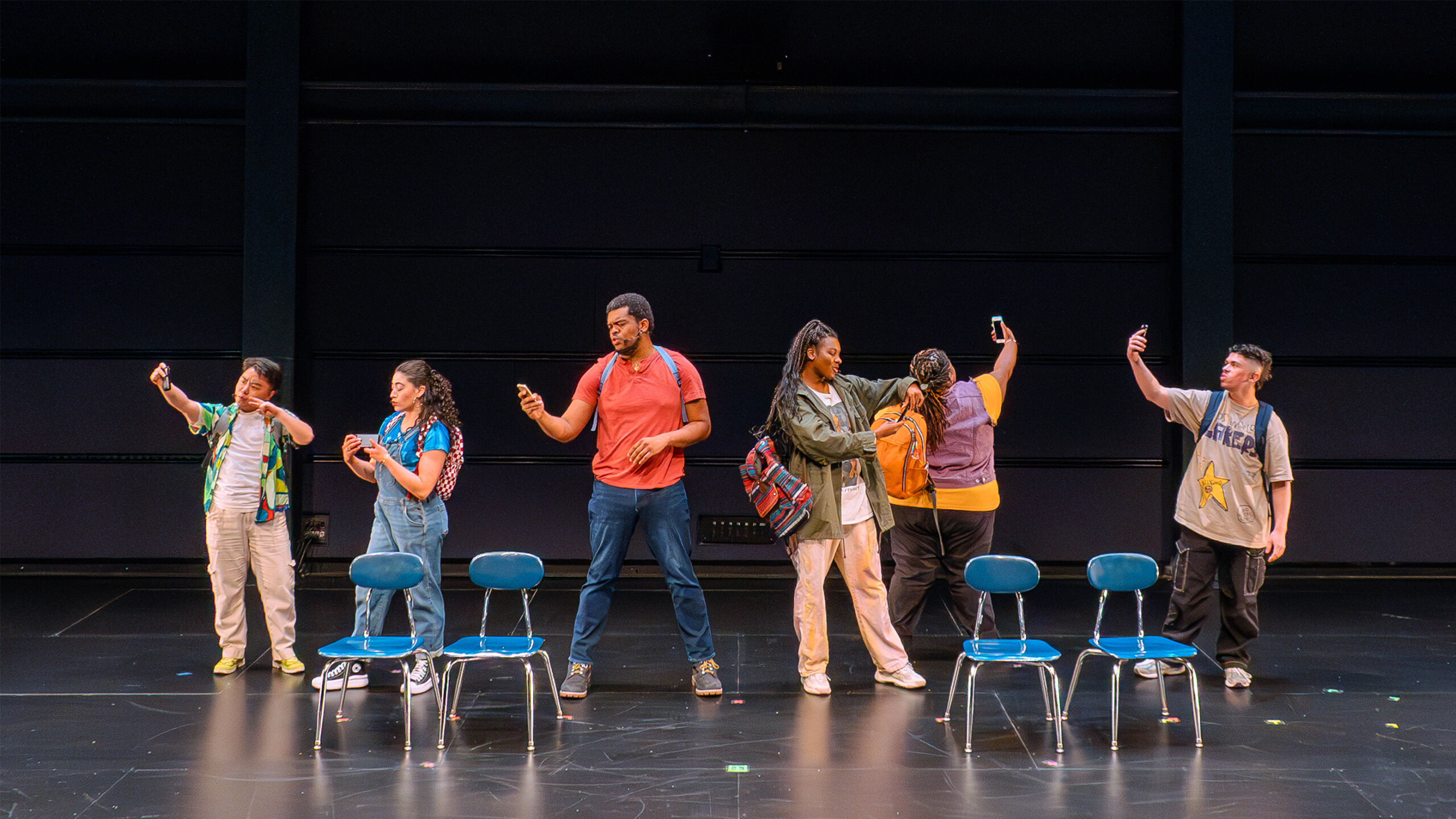 A diverse group of six individuals stands on a stage, each holding a smartphone to take selfies. They are arranged in a line with four blue chairs placed in front...