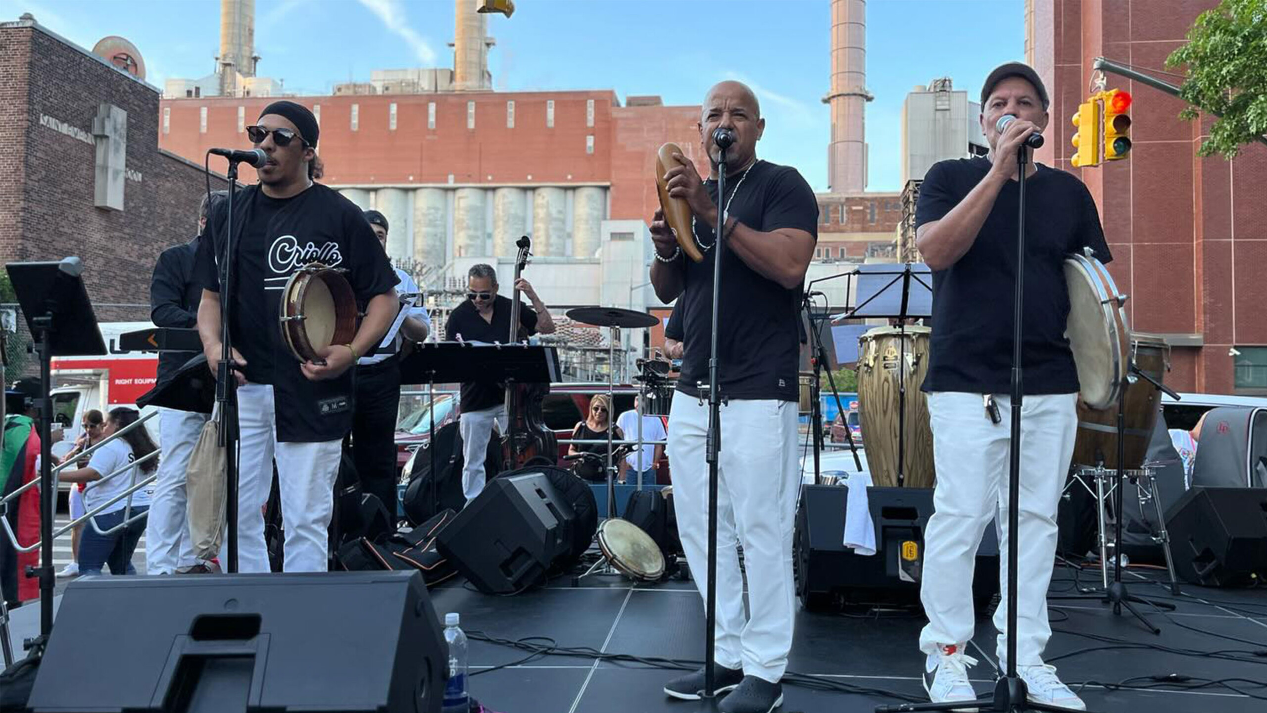 A lively band performs on a temporary outdoor stage in an urban environment. Three male musicians are prominently featured in the foreground, each dressed in a...