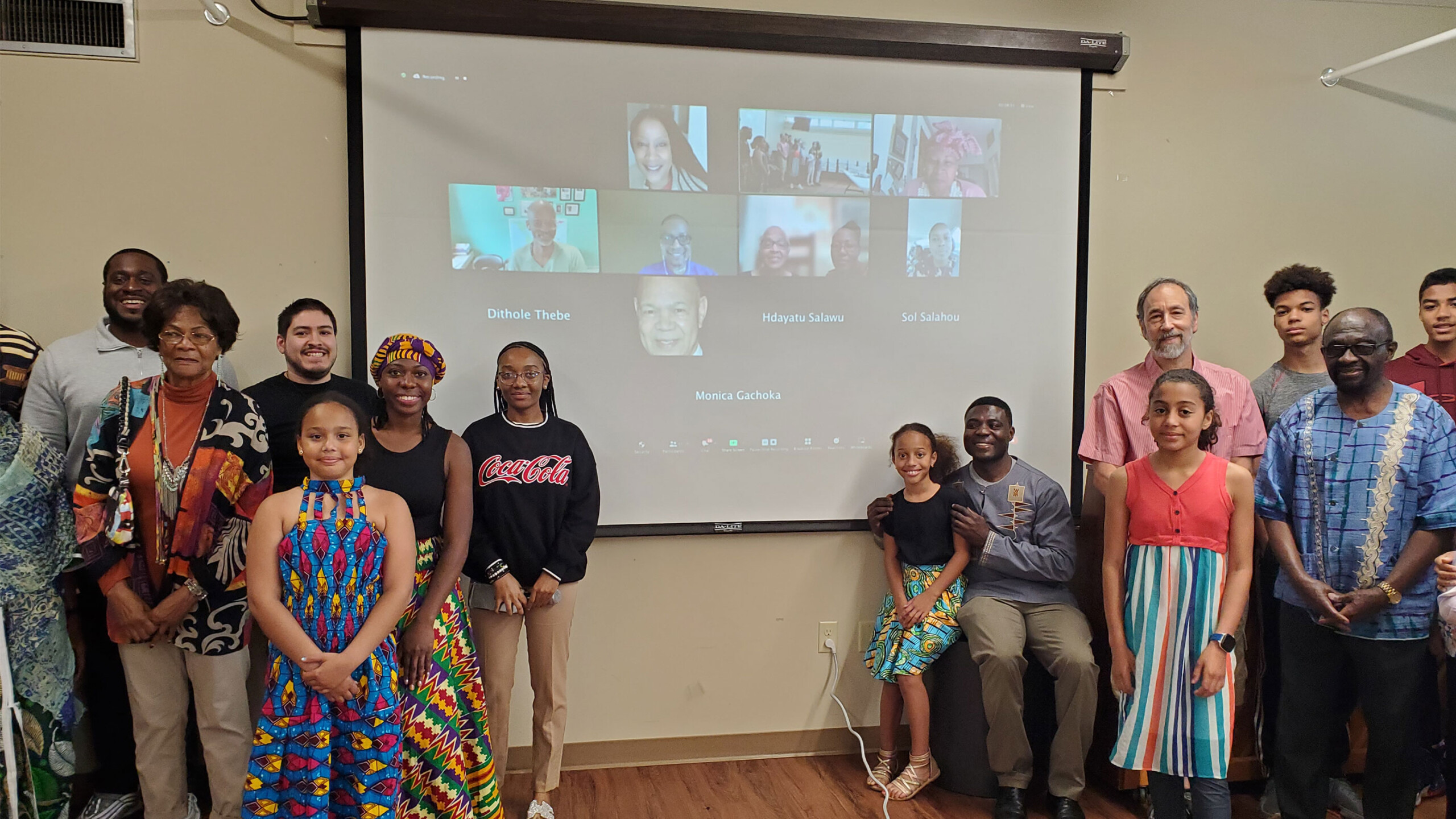 A diverse group of people of various ages stands in front of a large screen displaying a virtual meeting. The screen shows multiple participants in a video call,...