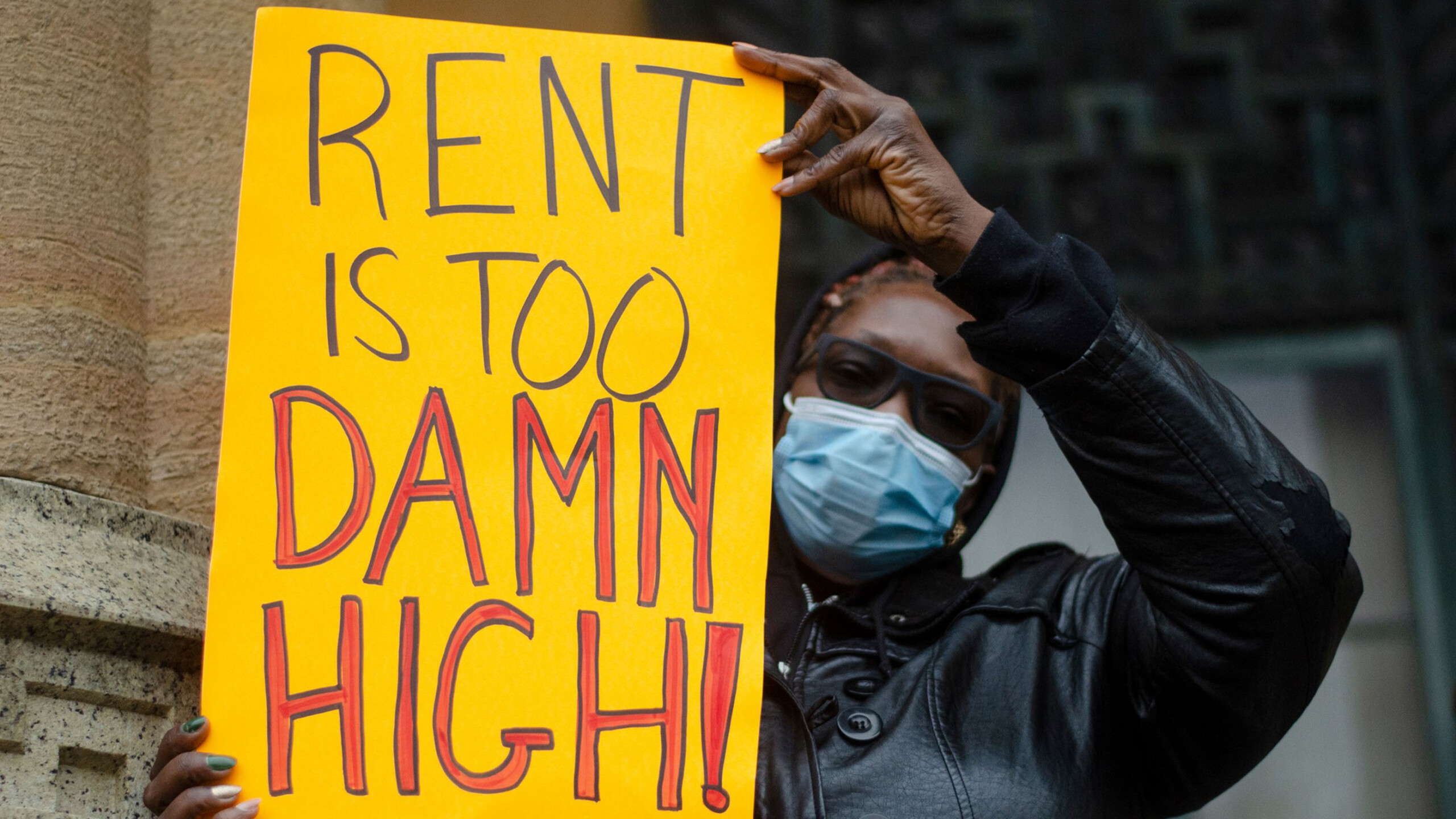 An individual protests against high rental costs, holding a bright yellow sign with bold black and red lettering that reads 'Rent is too damn high!' The person...