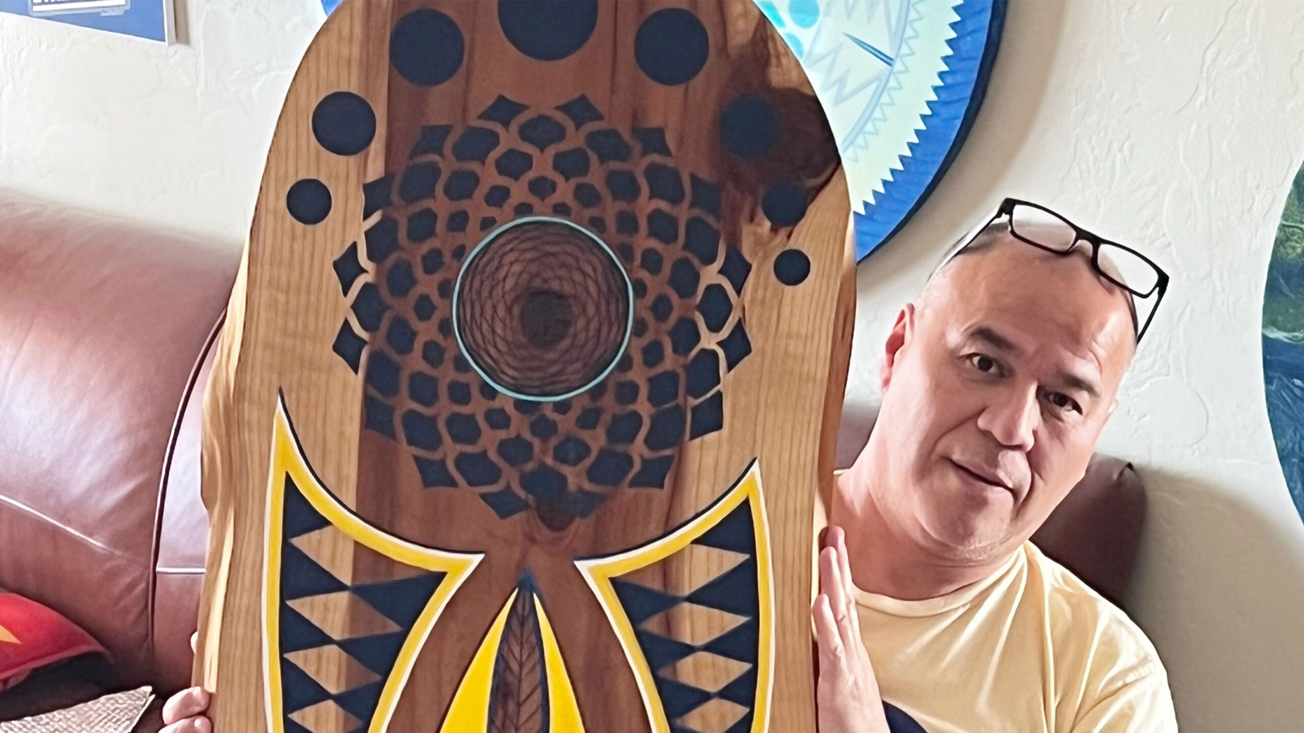 A man sitting on a brown leather couch is holding a wooden artwork with intricate geometric designs and bold colors. The artwork features a central circular pattern...