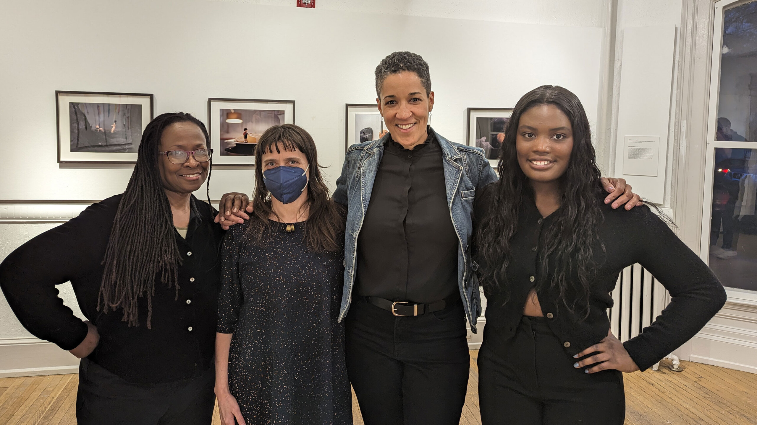 A group of people is posing for a photo indoors, likely in an art gallery. They are smiling and standing together, showcasing their clothing and expressions. The...