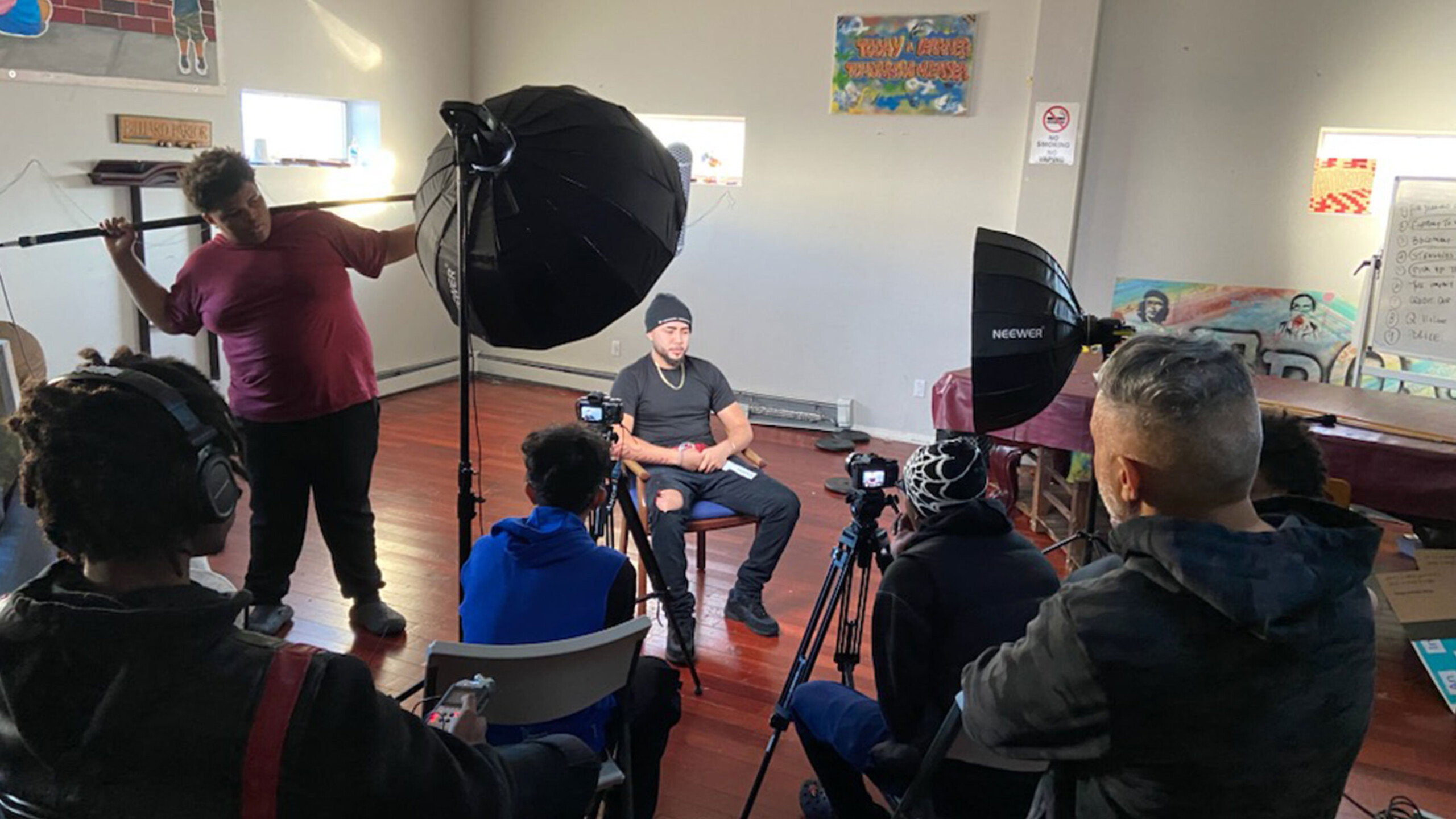 A group of production crew members is setting up for an interview. A man sits on a chair in the center, facing a camera. He appears to be the interviewee, dressed...