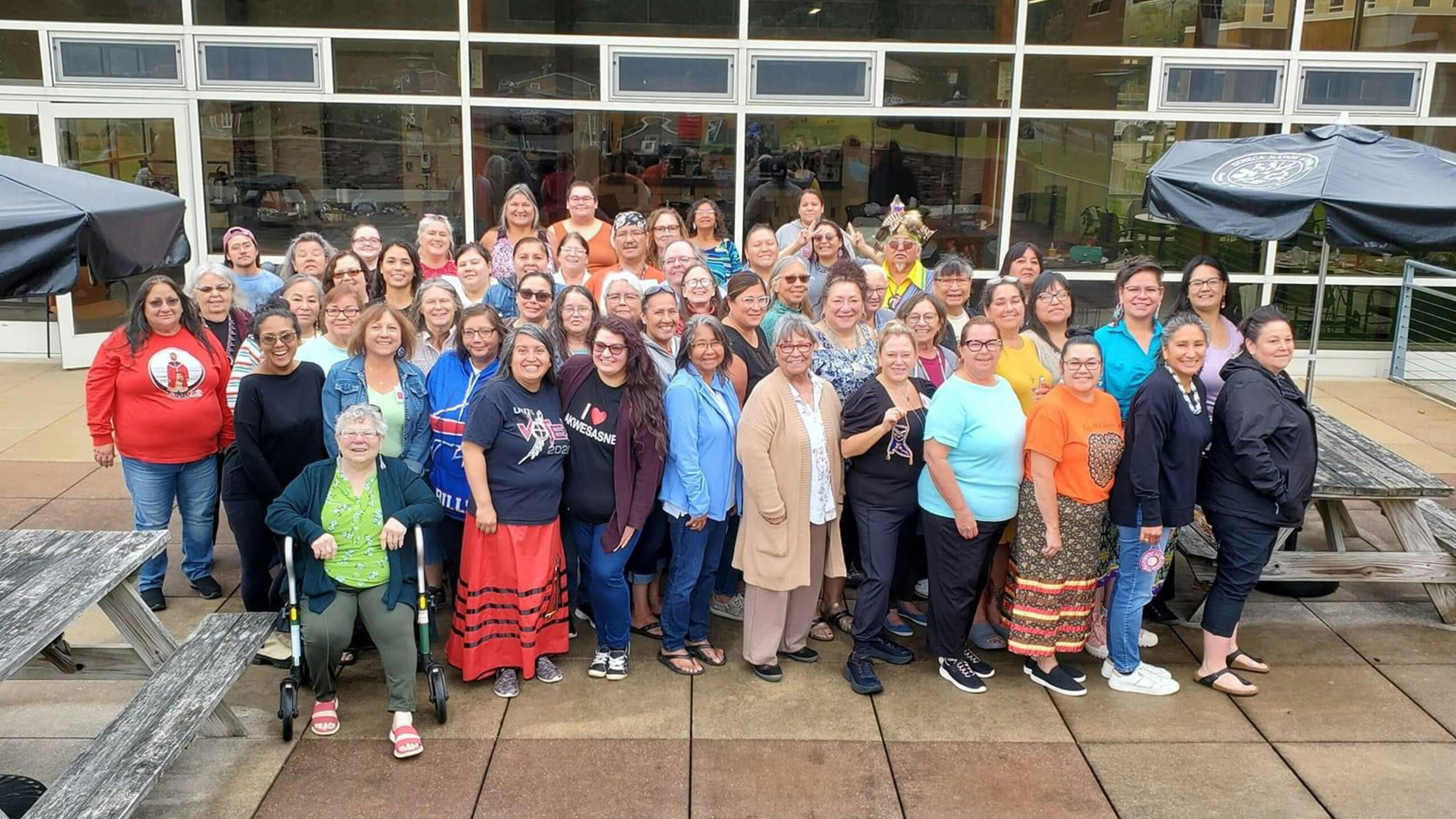 A large group of diverse individuals, mostly women, gather closely together for a photo. They are standing outdoors on a paved area with picnic tables nearby. Many...