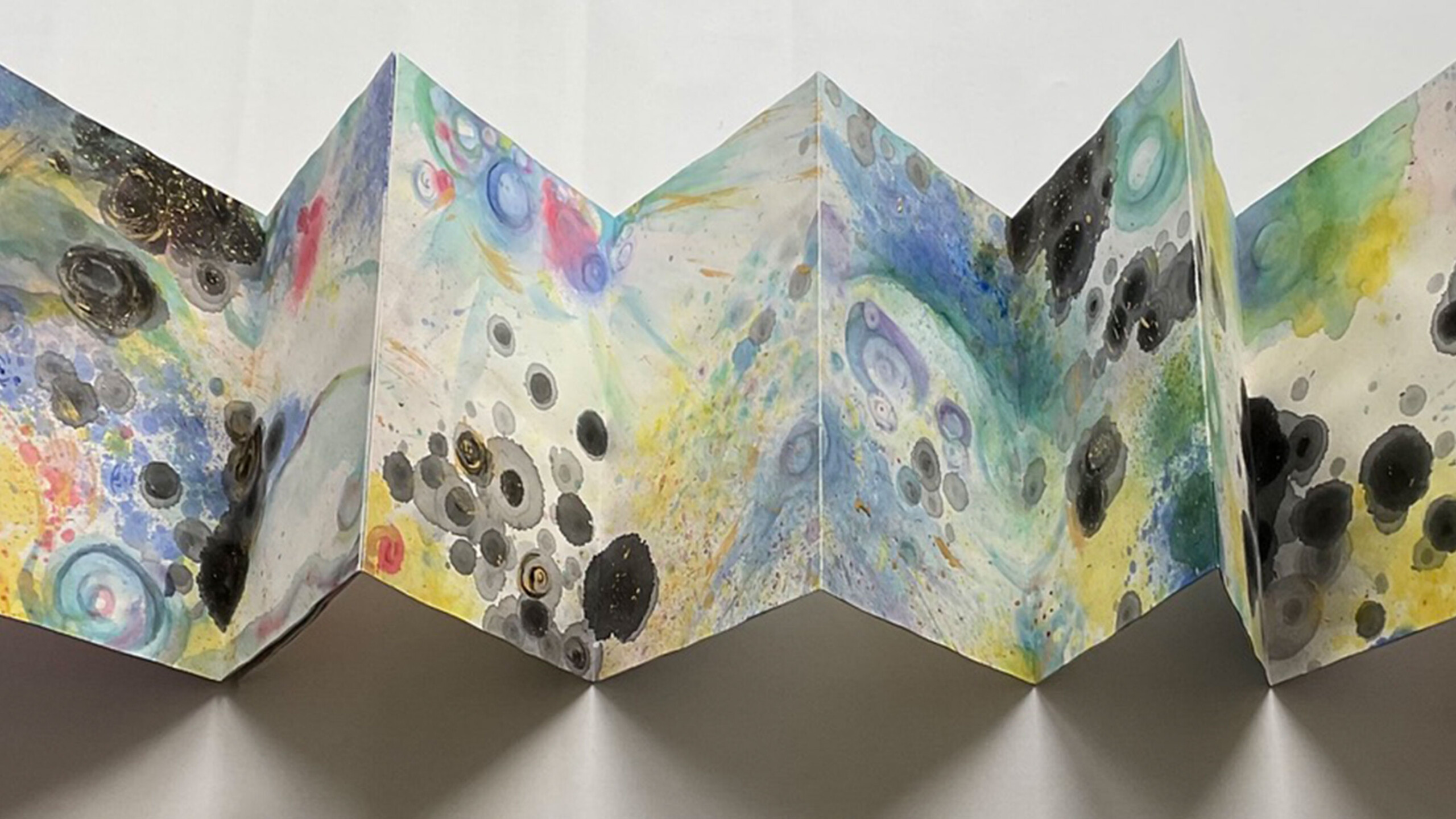 A vivid abstract artwork displayed on an accordion-style folded paper. Bright and dark colors blend across the panels, featuring swirling patterns of blues, yellows,...